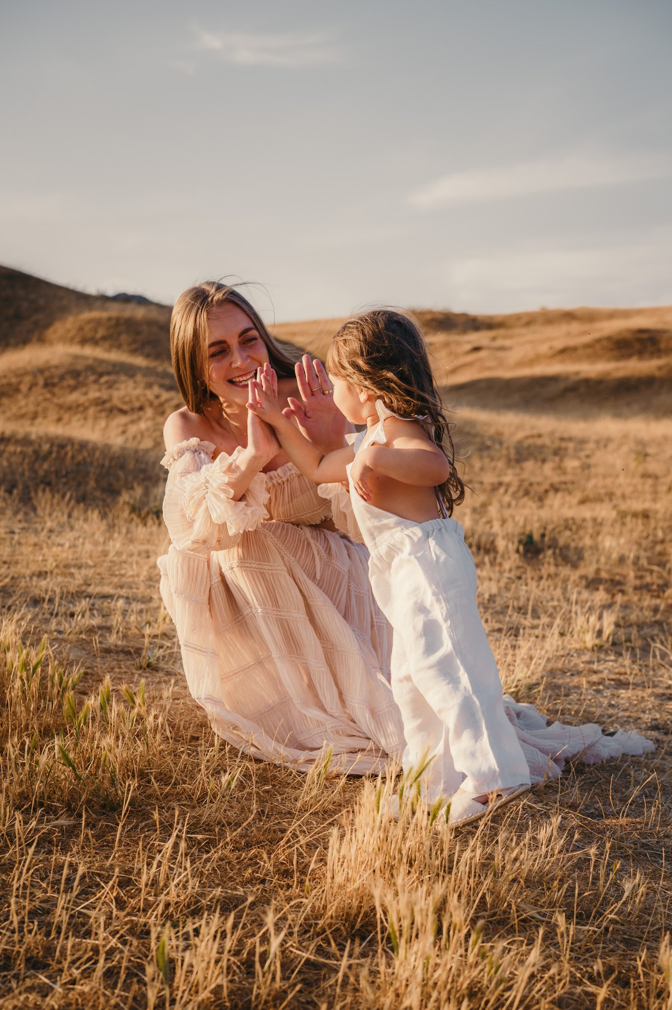 A woman and a young girl with brown hair playing together outdoors in a grassy, hilly landscape during sunset, facing each other and touching hands.