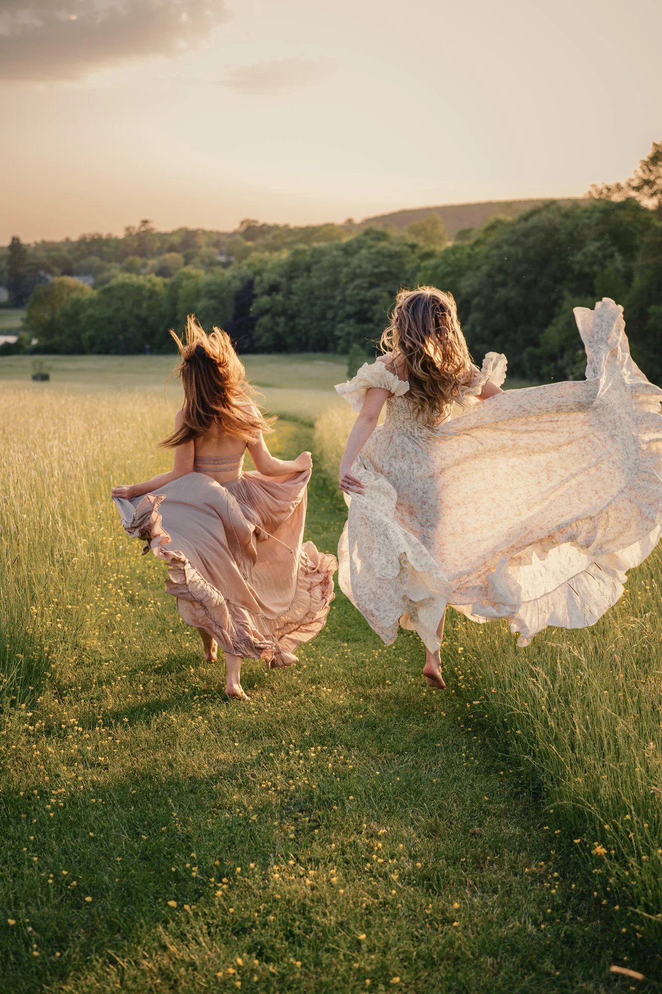 Two women in vintage dresses running barefoot through a grassy field during sunset.