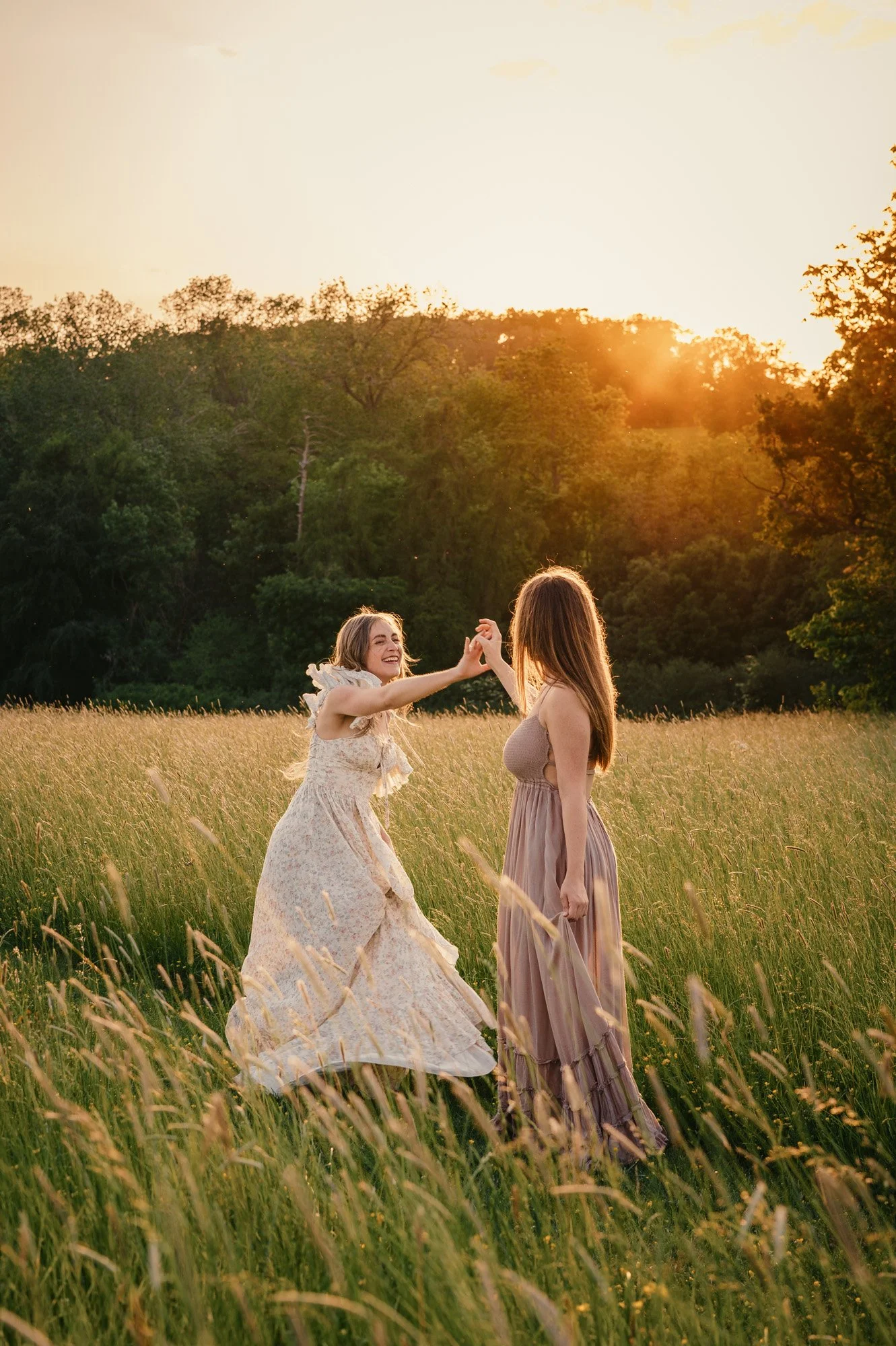 Two women dancing in a field of tall grass during sunset, surrounded by trees.