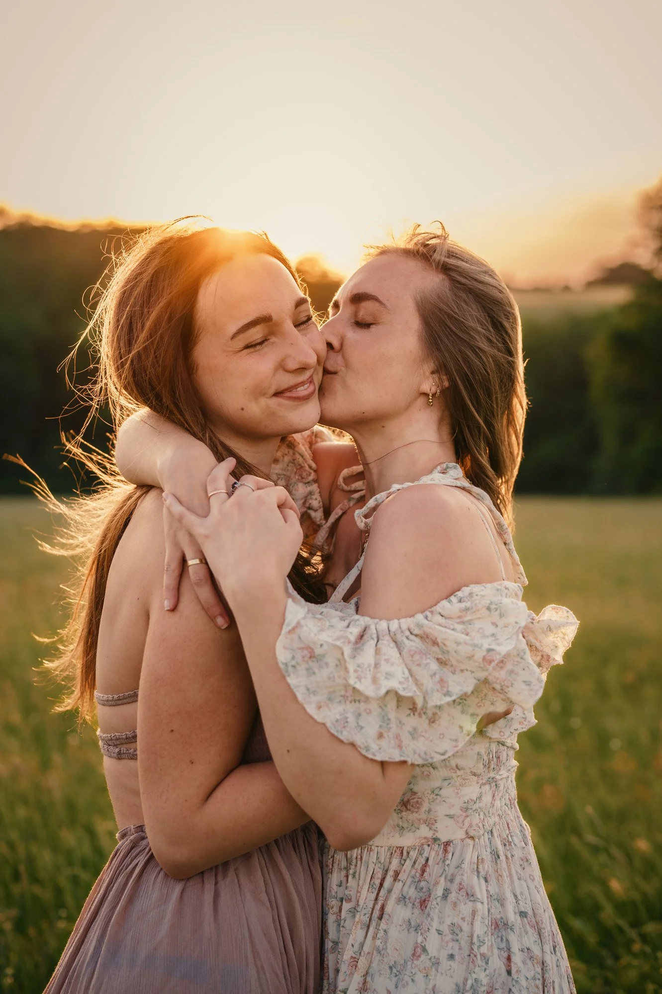 Two women embrace outdoors at sunset, one kissing the other on the cheek, both smiling with eyes closed. They are holding each other in a field with trees and a glowing sky in the background.