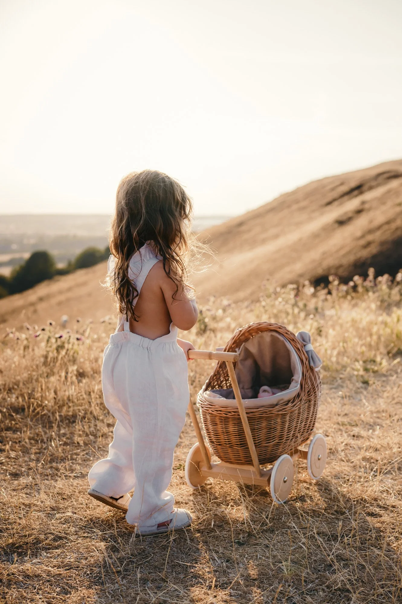 A young girl with long, curly hair stands outdoors in a grassy field during sunset, pushing a wicker baby stroller with a blanket inside.