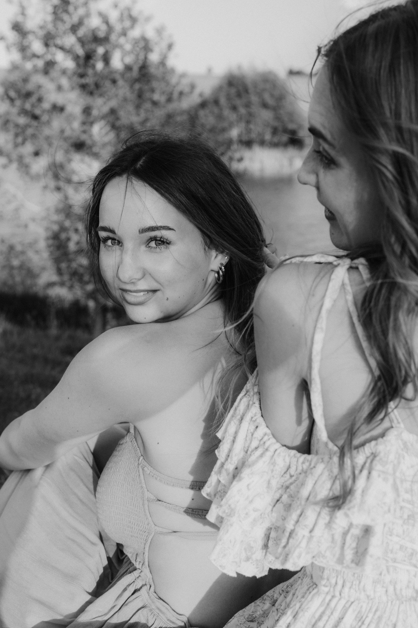Two women sitting outdoors near a body of water with trees in the background, smiling and looking at each other, one with dark hair and the other with light hair, in black and white.