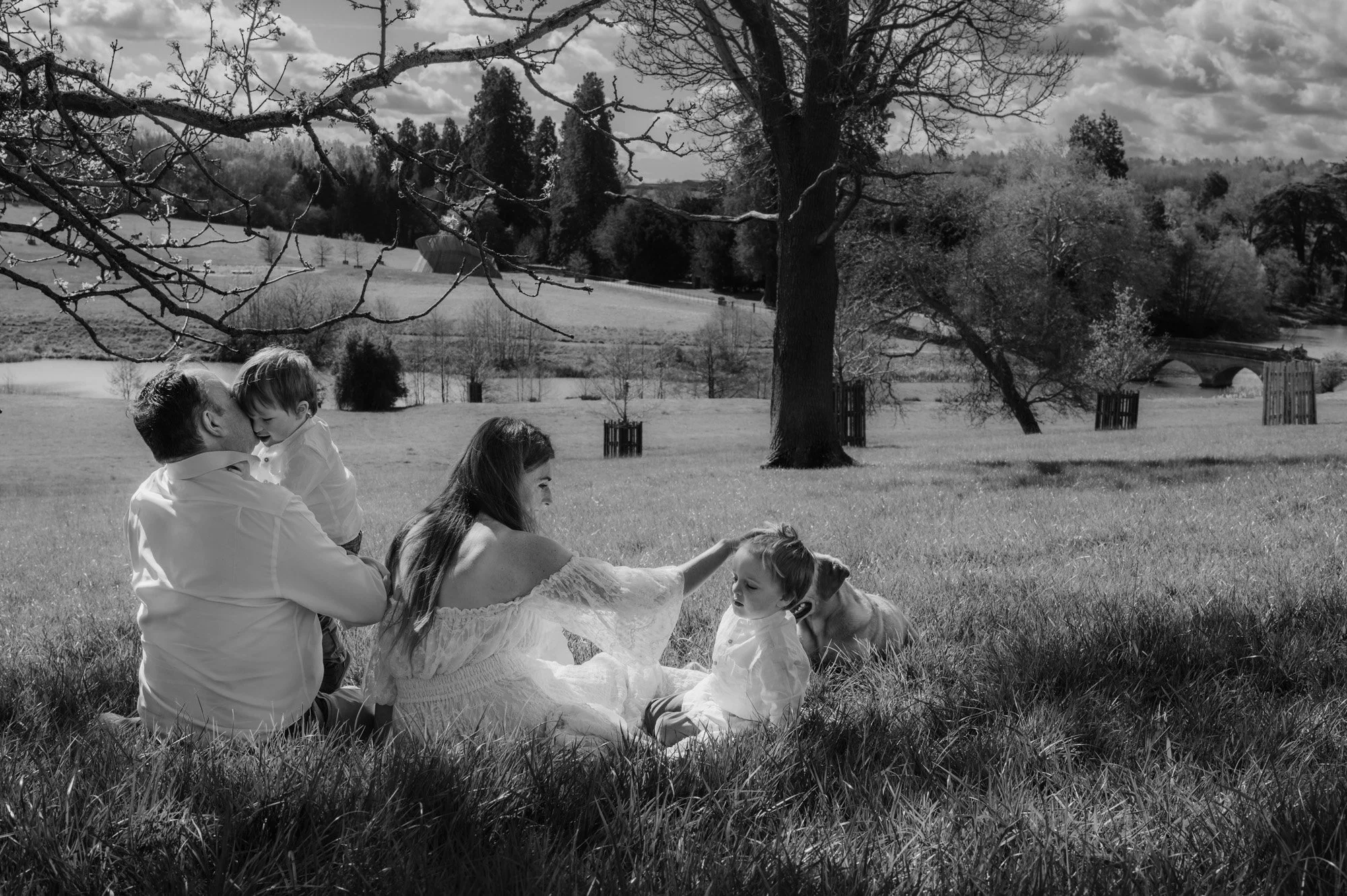 Black and white photo of a family in a park, including two children and two adults. The man is holding a boy, and the woman is petting a girl. Trees and a bridge are visible in the background.