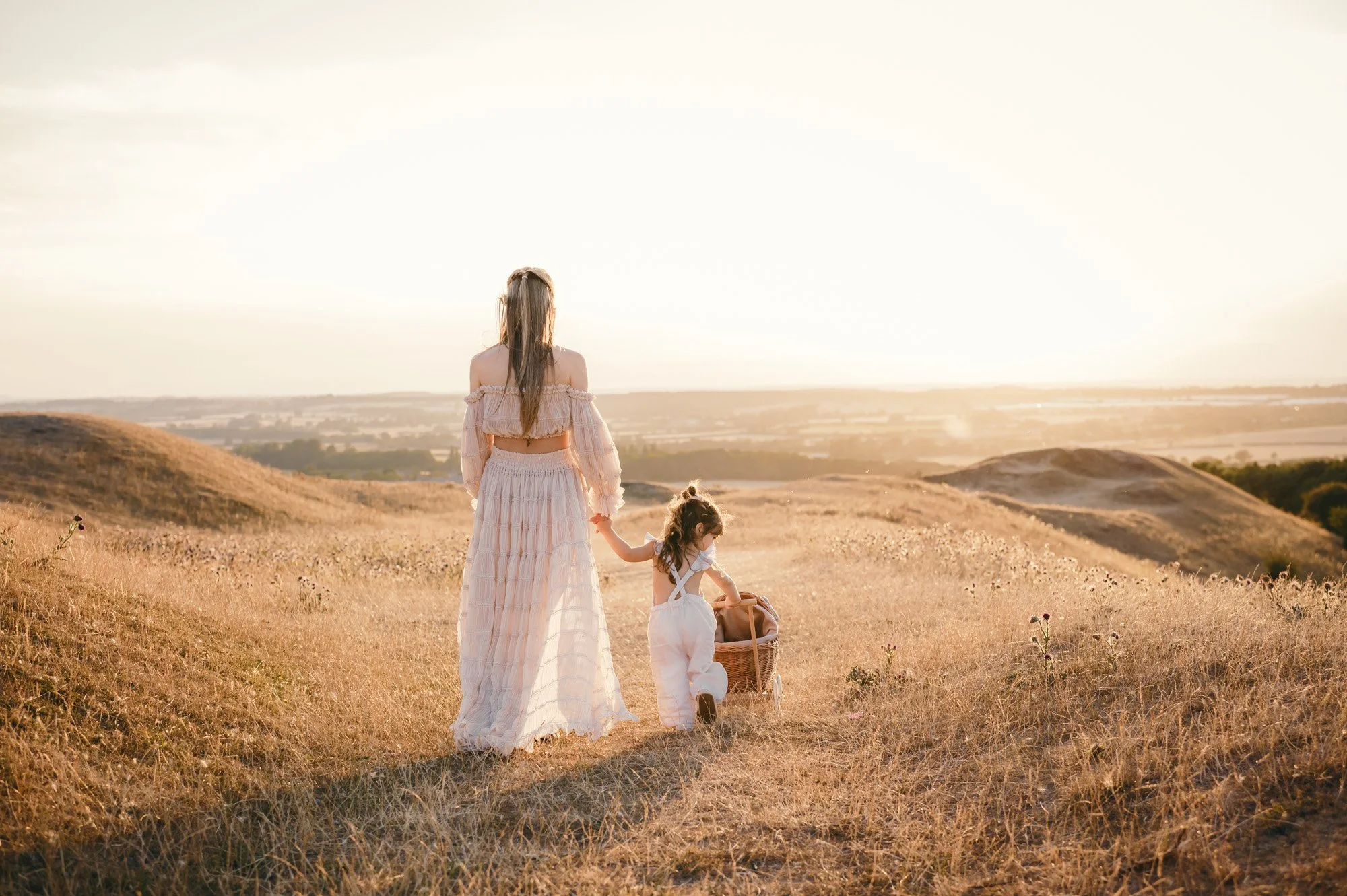 A woman and two young girls walking through a golden field at sunset, with rolling hills and a distant cityscape in the background.