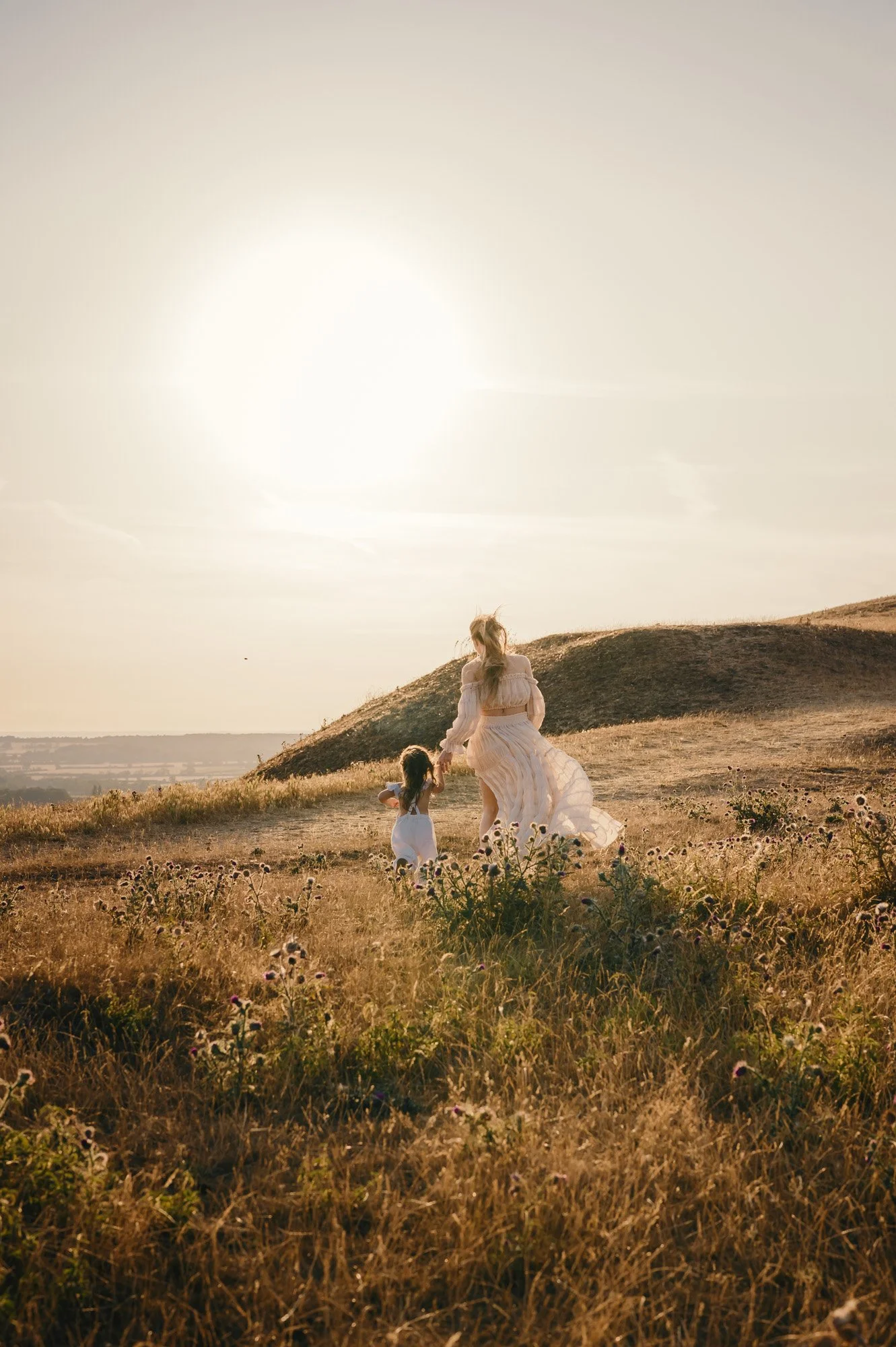 A woman in a flowing white dress walking hand-in-hand with two young girls through a grassy field at sunset, with hills in the background.