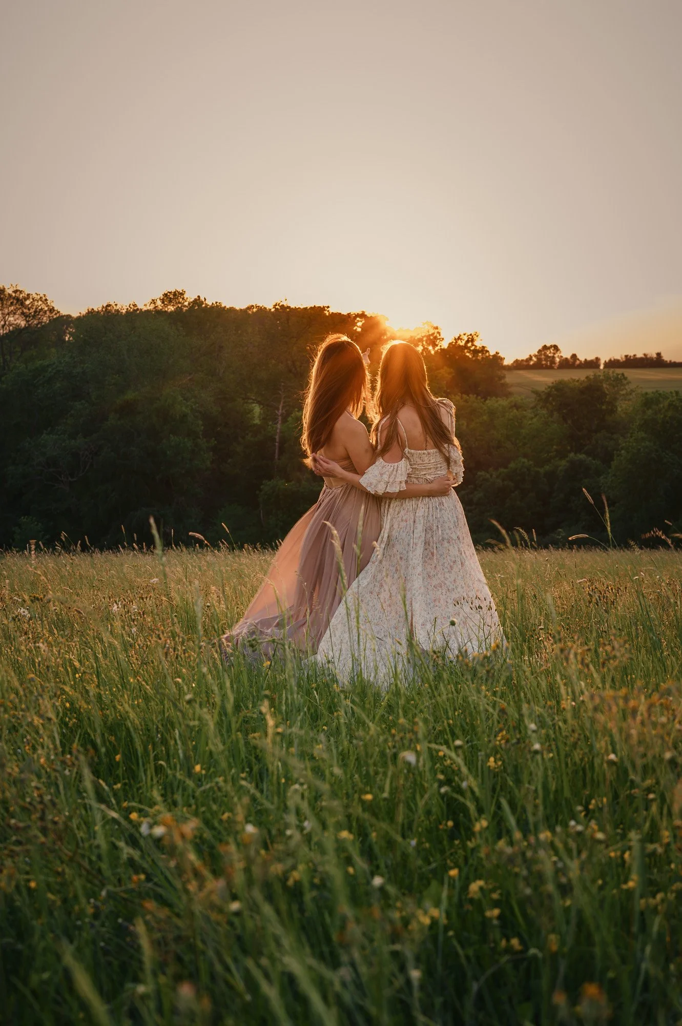 Two women with long hair, wearing flowing dresses, embracing each other in a grassy field at sunset with trees in the background.