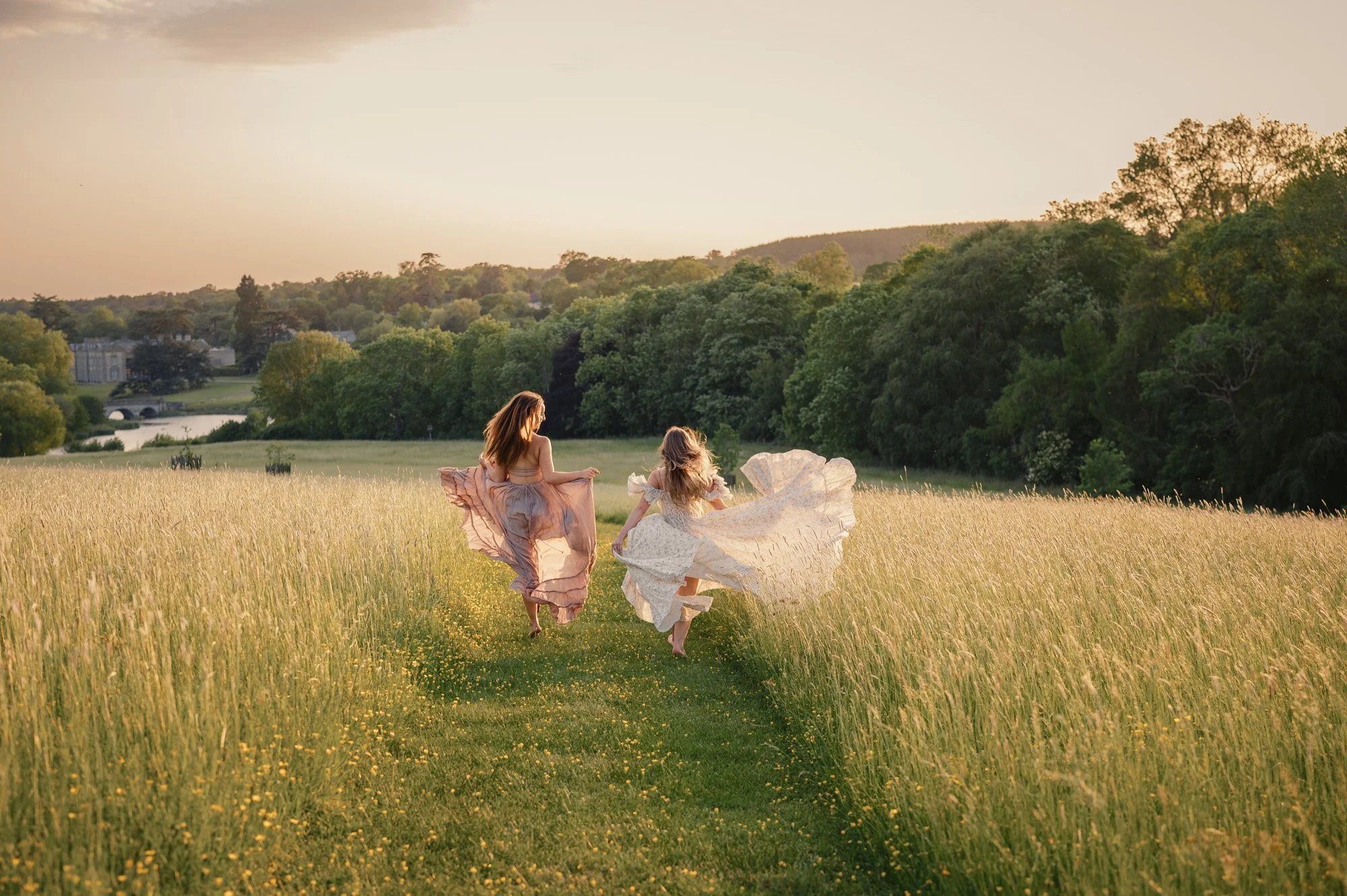 Two women in flowing dresses running through a yellow wildflower field at sunset with lush green trees and a distant bridge in the background.