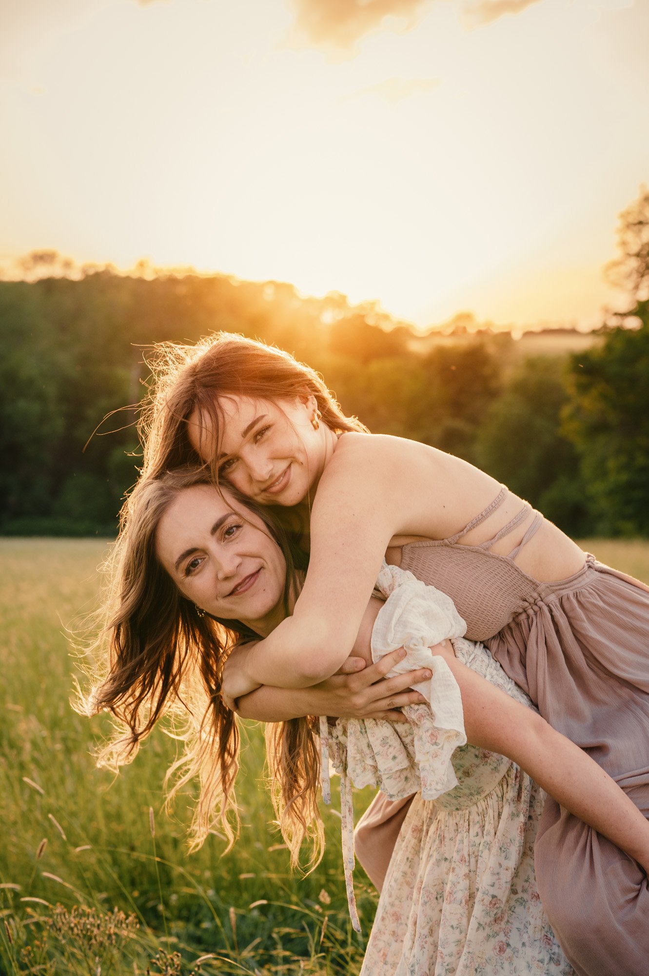 Two women are smiling and embracing each other outdoors during sunset, with a field and trees in the background.