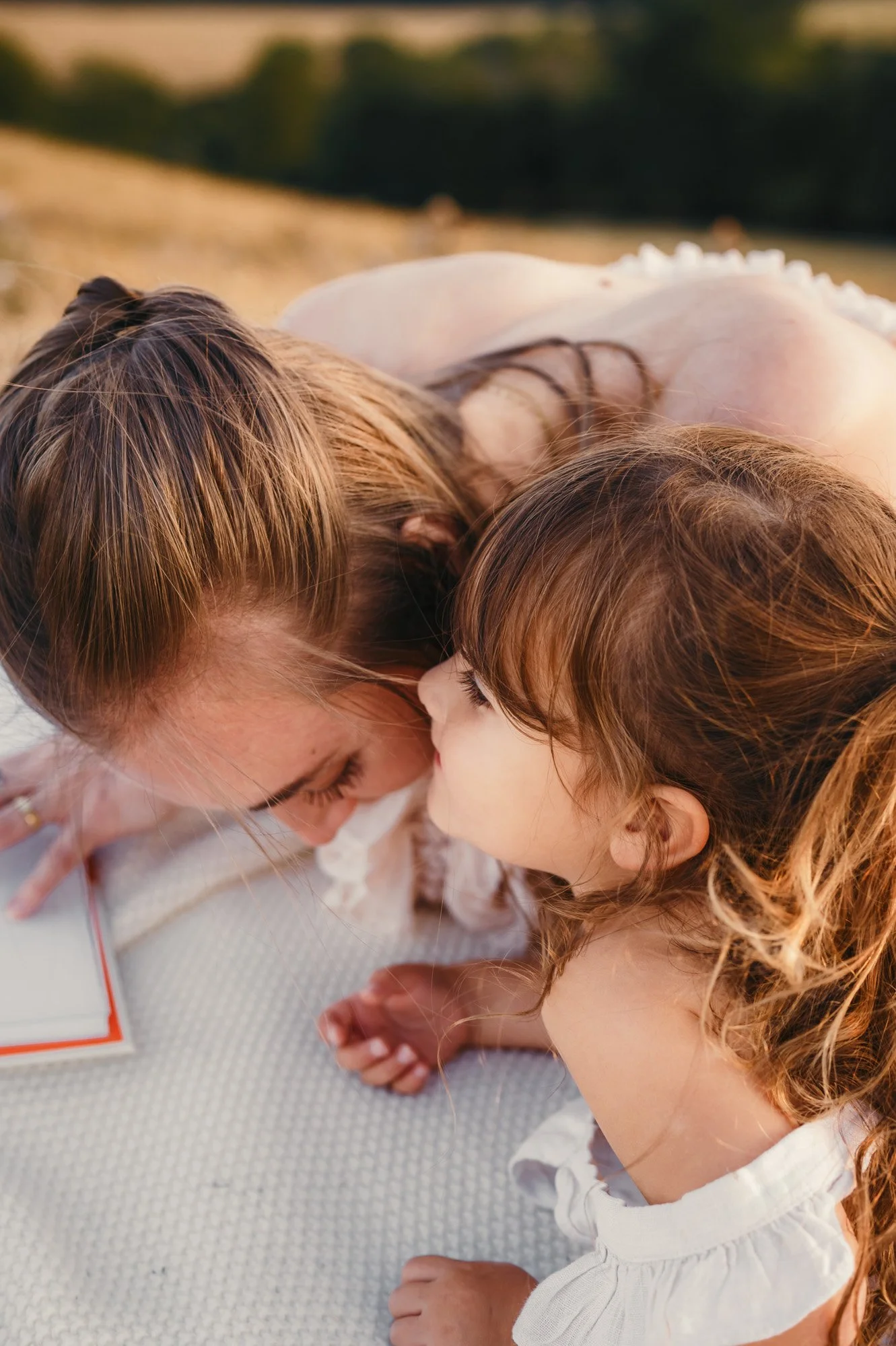 A woman and a young girl are lying face to face on a light-colored textured surface, with their foreheads touching and eyes closed, appearing to share a tender moment.