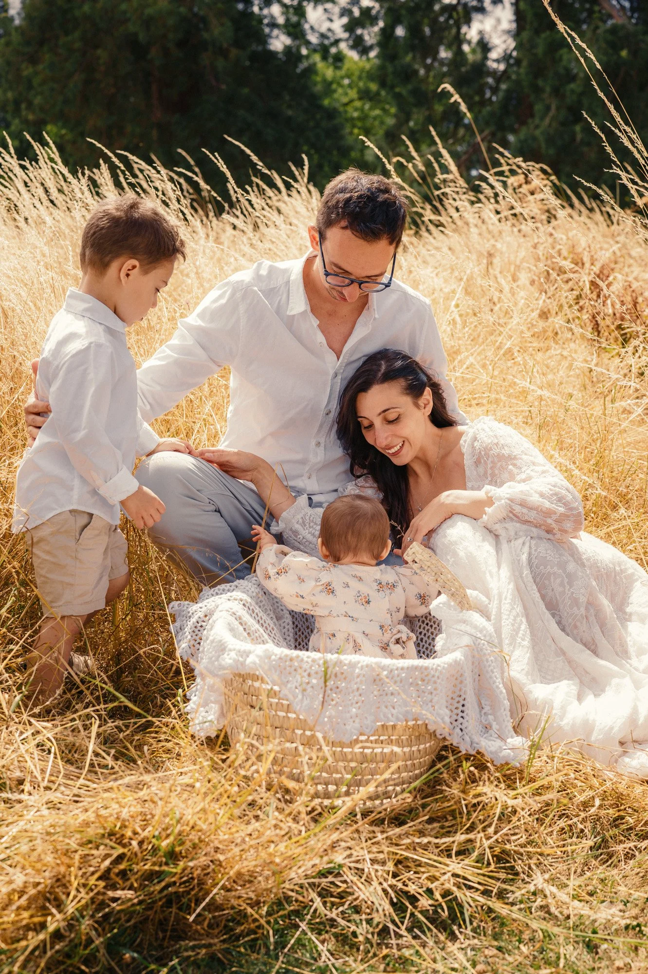 Family of four, including a mother, father, and two young children, sitting in a golden field of tall grass, with a wicker basket in the foreground and trees in the background, enjoying a picnic in the late afternoon.