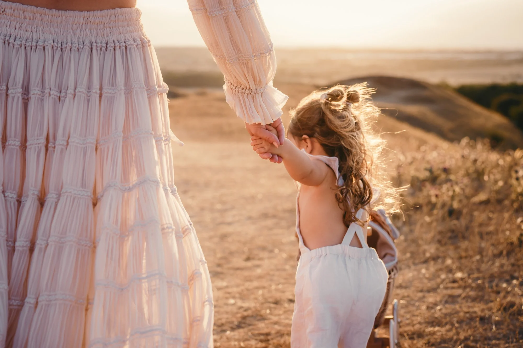 A woman and a little girl holding hands on a beach during sunset. The woman is wearing a pink, flowing, long-sleeved dress, and the girl is dressed in a white outfit. The focus is on their hands and the soft sunlight enhances the warm, peaceful atmos