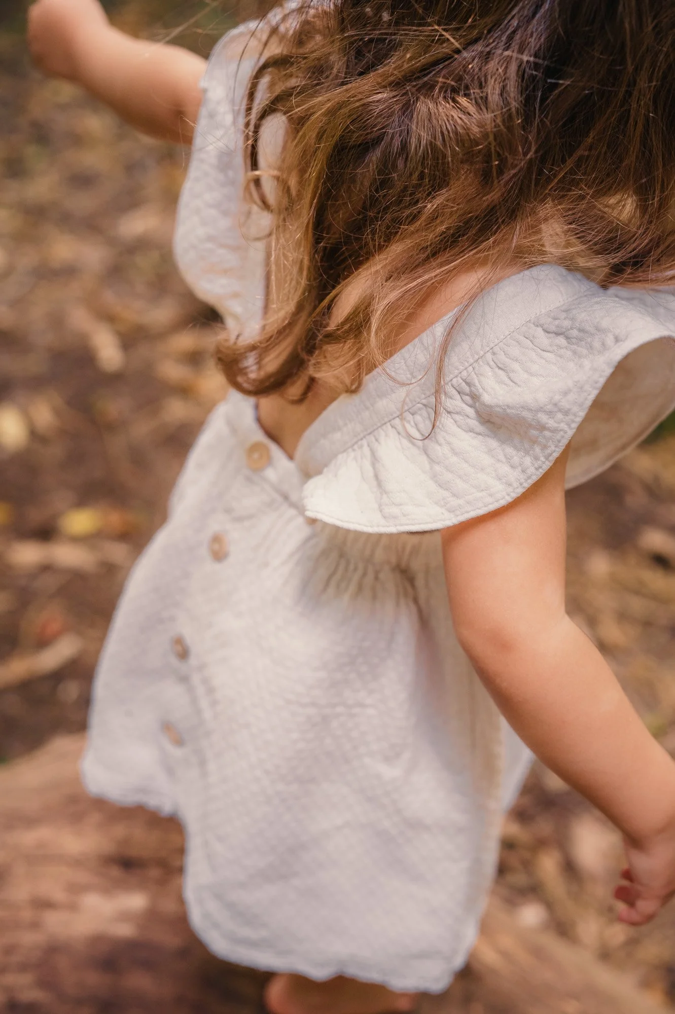 Close-up of a young girl with light brown hair, wearing a light-colored dress with buttons, standing outdoors on a dirt path.