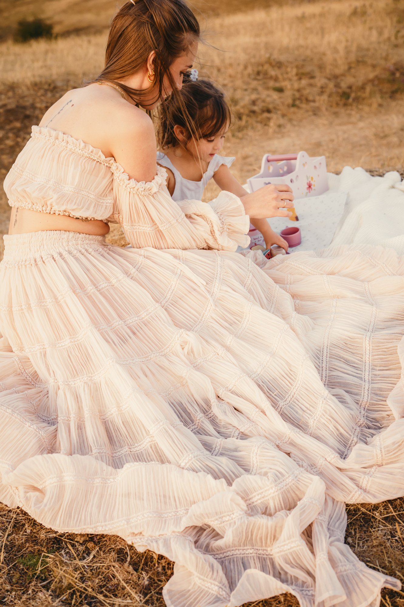 A woman in a peach-colored off-shoulder dress sitting on dry grass with a young girl, playing with small toys and a box nearby. The scene is outdoors during sunset.