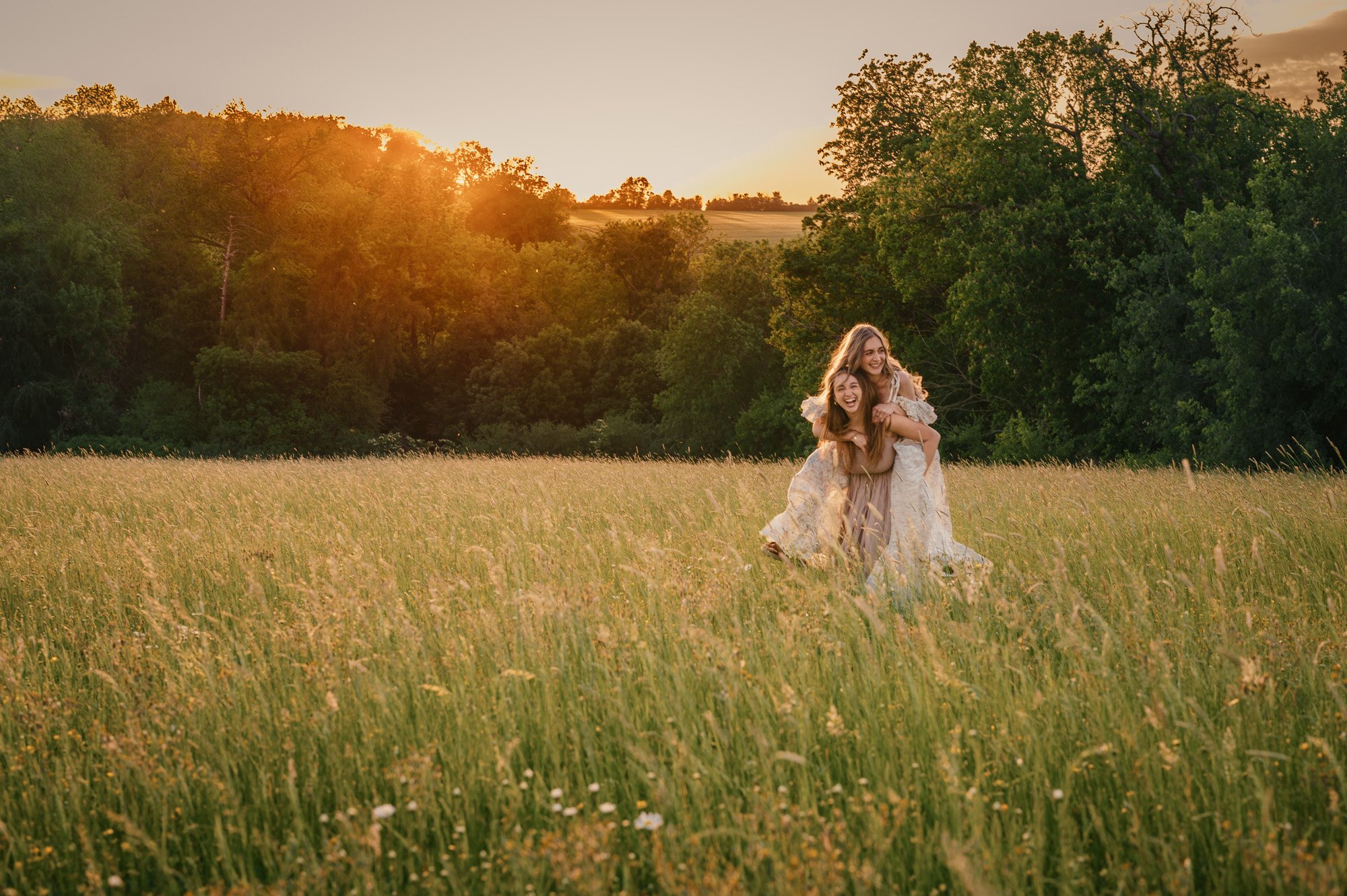 Two women in white dresses are playing and laughing in a field of tall grass during sunset, surrounded by trees.