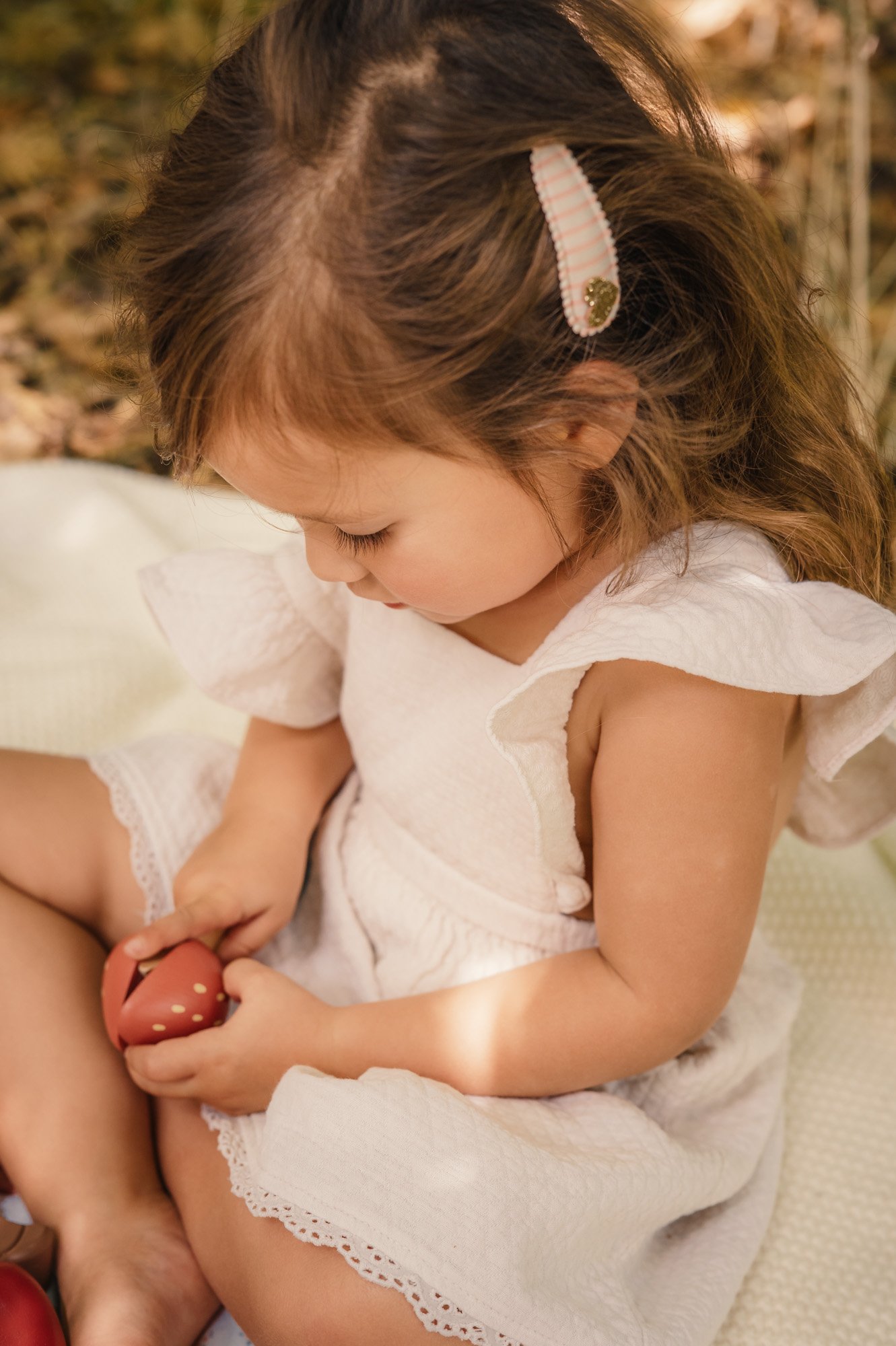 A young girl with brown hair clipped with a pink and white striped hair clip with a gold decoration, wearing a white dress with lace trim, sitting down and looking at a strawberry in her hands.