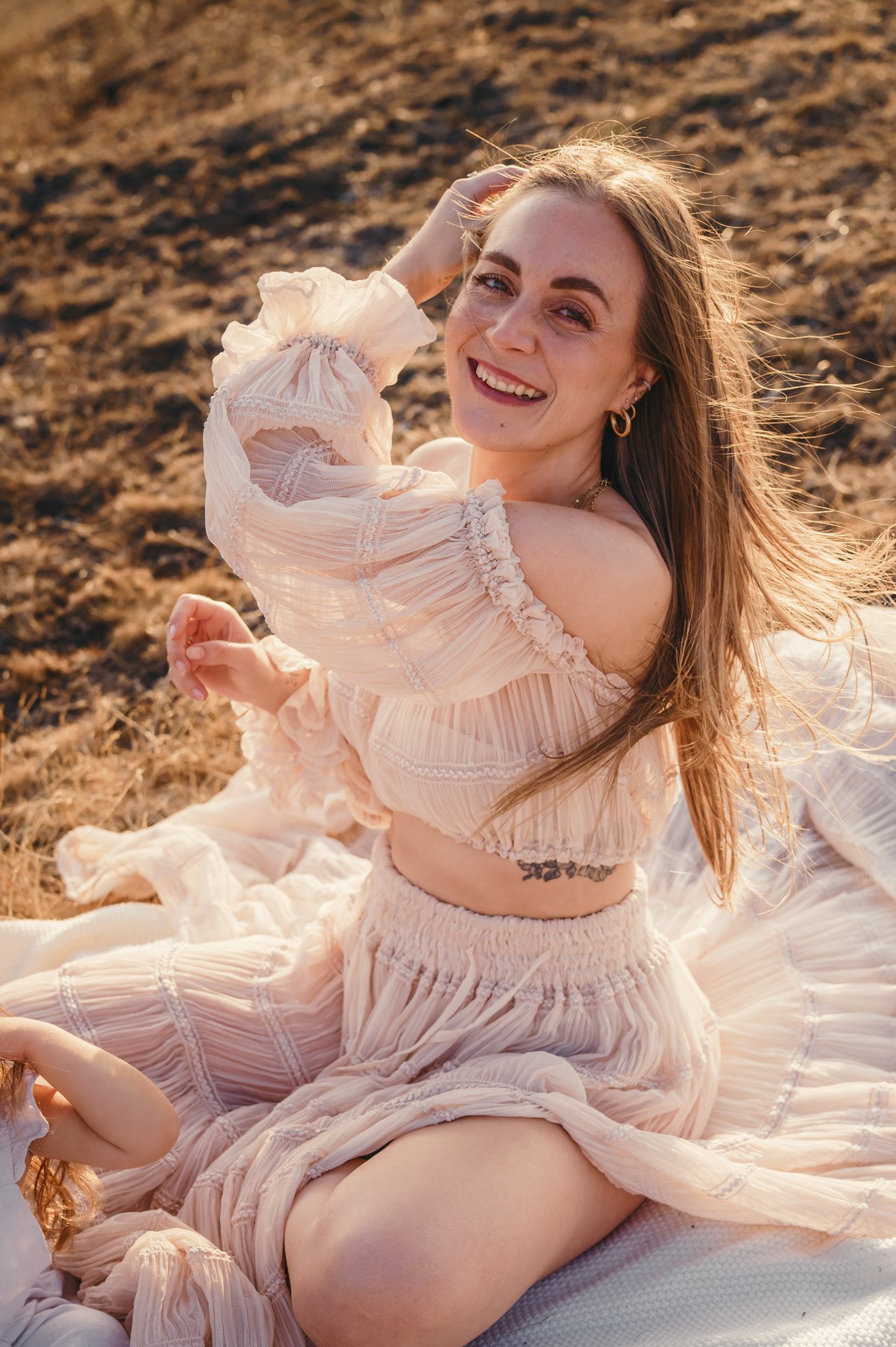 A smiling woman with long hair and gold hoop earrings, wearing a light pink, ruffled dress, sitting outdoors on a blanket in a sandy area.