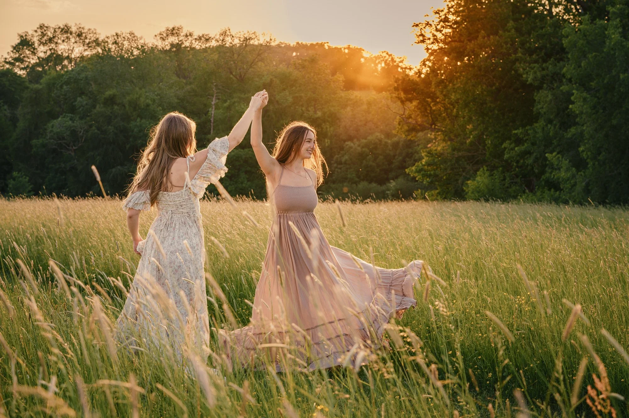 Two women in long dresses holding hands and dancing in a meadow during sunset with green trees in the background.