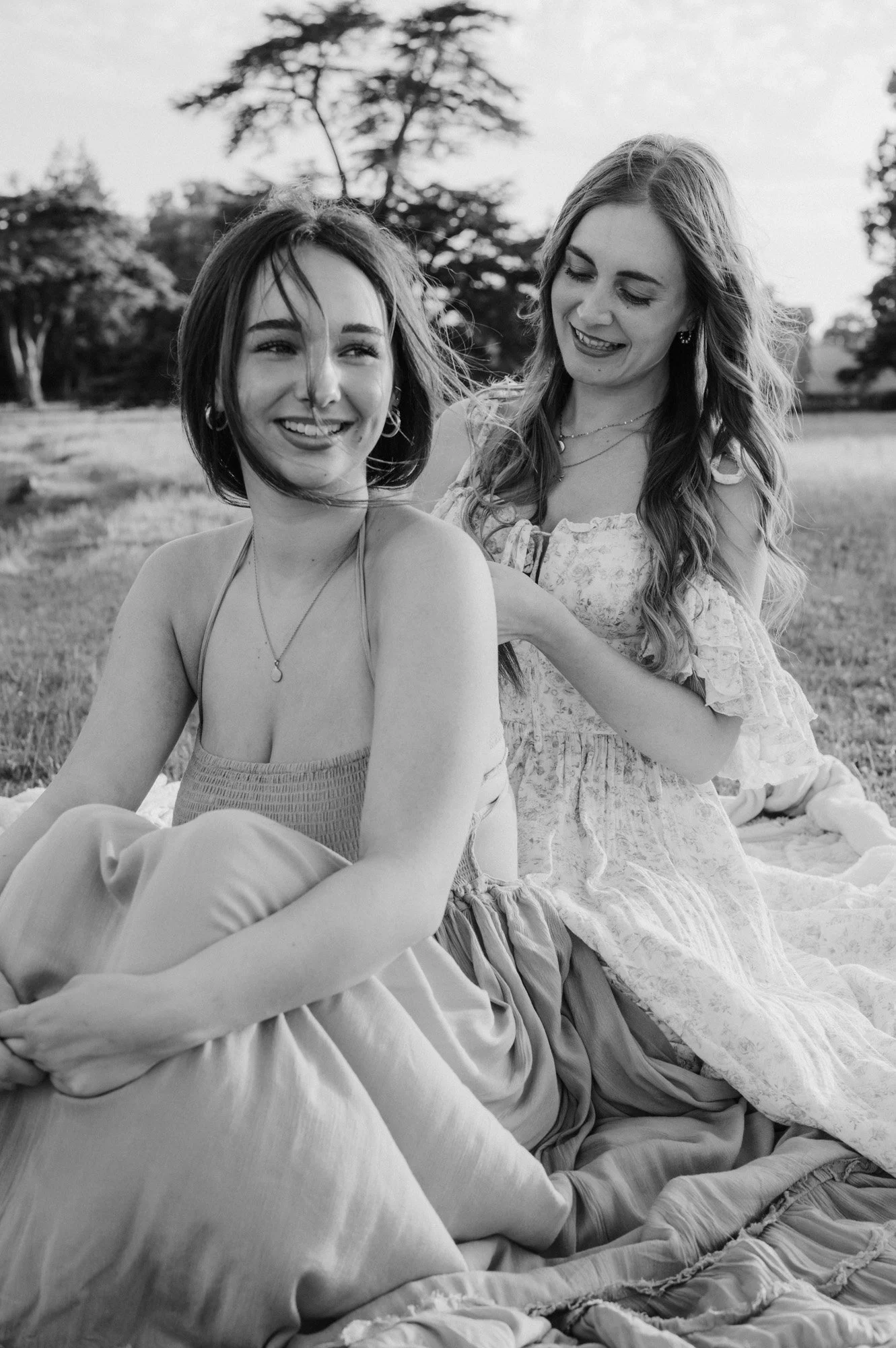 Two women sitting outdoors in a grassy area, smiling and enjoying each other's company. One woman has short hair and is wearing a spaghetti strap top, while the other has long hair and is wearing a dress with ruffled sleeves. There are trees in the b