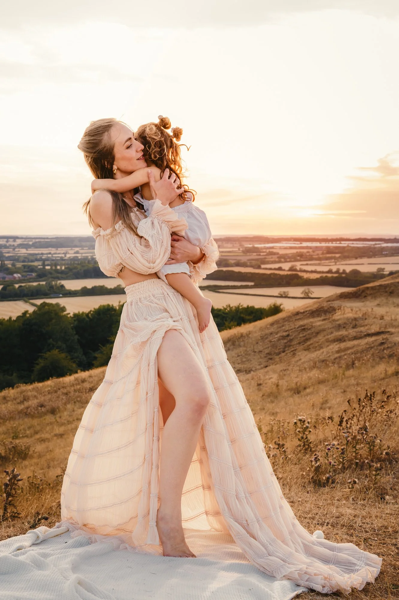 A woman in a flowing peach-colored dress holding a young girl in an outdoor setting during sunset, with fields and a river in the background.