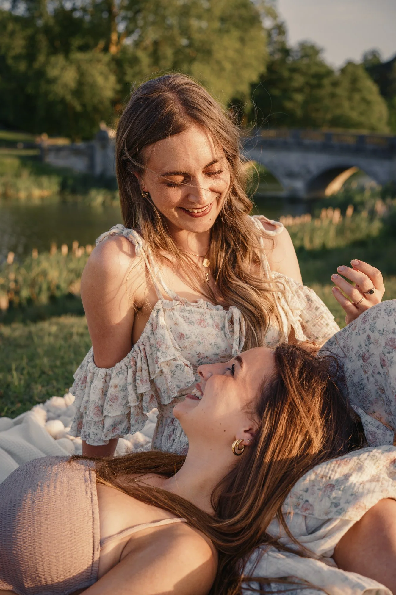 Two women lying on a blanket in a park, smiling and looking at each other, with a bridge and trees in the background during sunset.