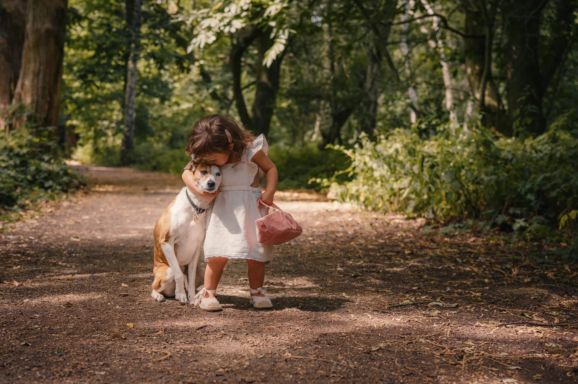 Young girl in white dress hugging a medium-sized dog with a white and brown coat on a dirt path in a forested area.