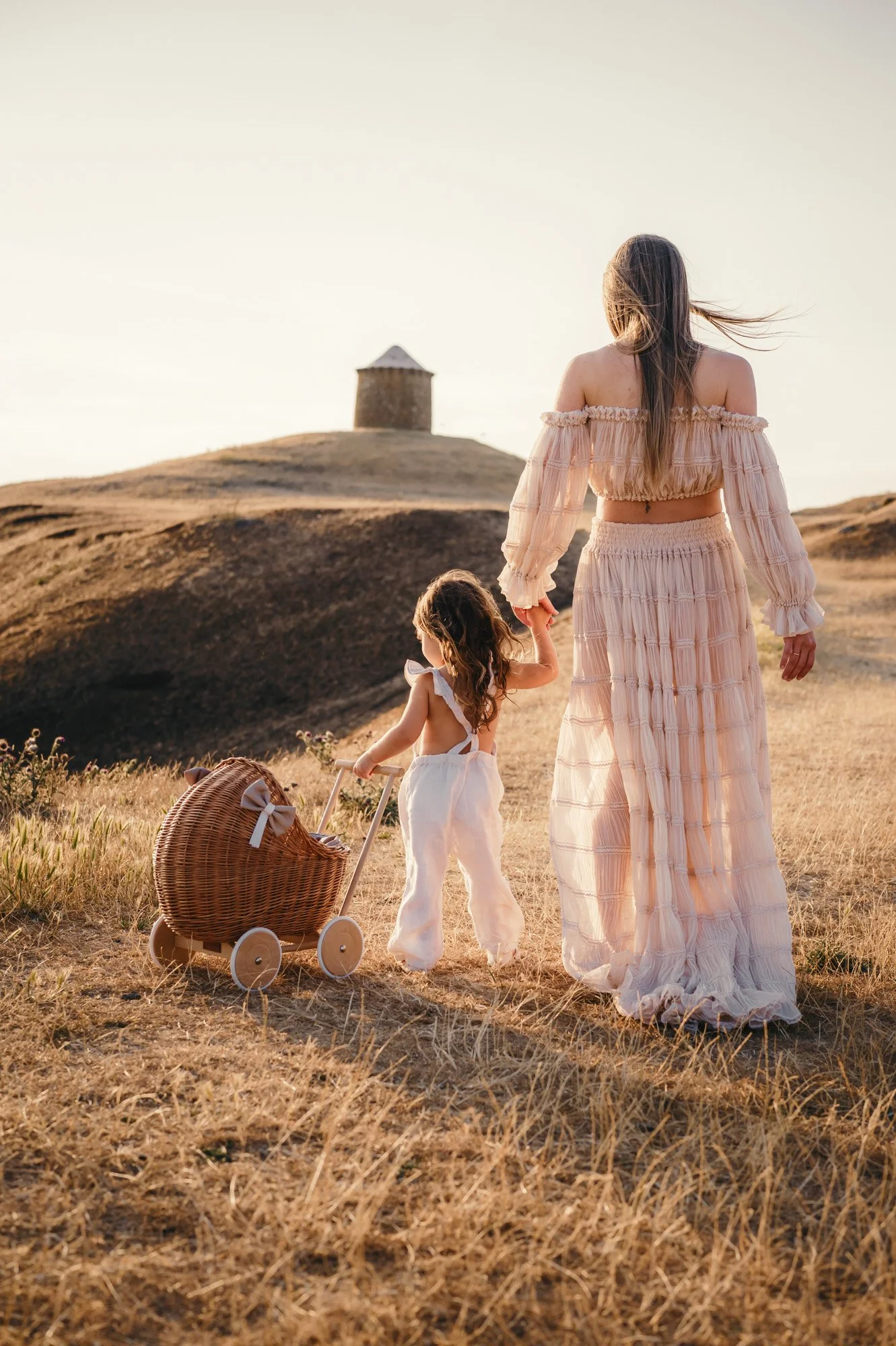 A woman and a young girl walk hand in hand through a golden field towards a small, stone tower on a hill in the distance at sunset. The woman wears a flowing, off-the-shoulder, peach-colored skirt and blouse, while the girl wears a white dress. A wicker stroller is on the ground beside them.