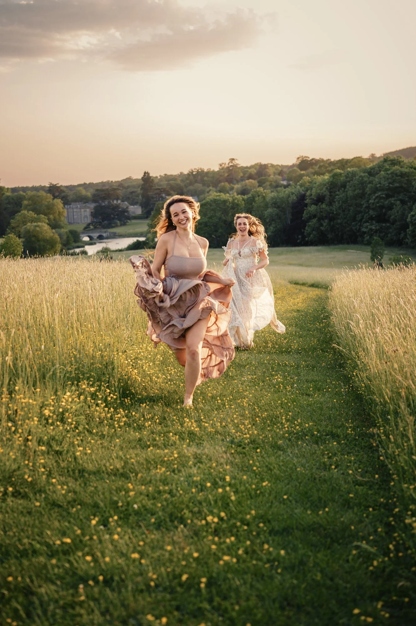Two women wearing elegant dresses running through a yellow flower field at sunset with a tree-filled landscape and a building in the background.