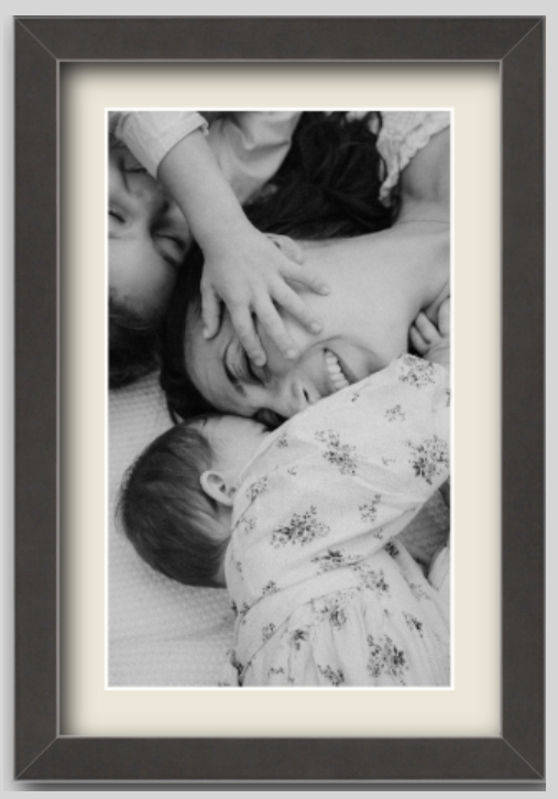 A black and white photo of a woman and two children lying on a bed, with the woman smiling and the children playfully touching her face.