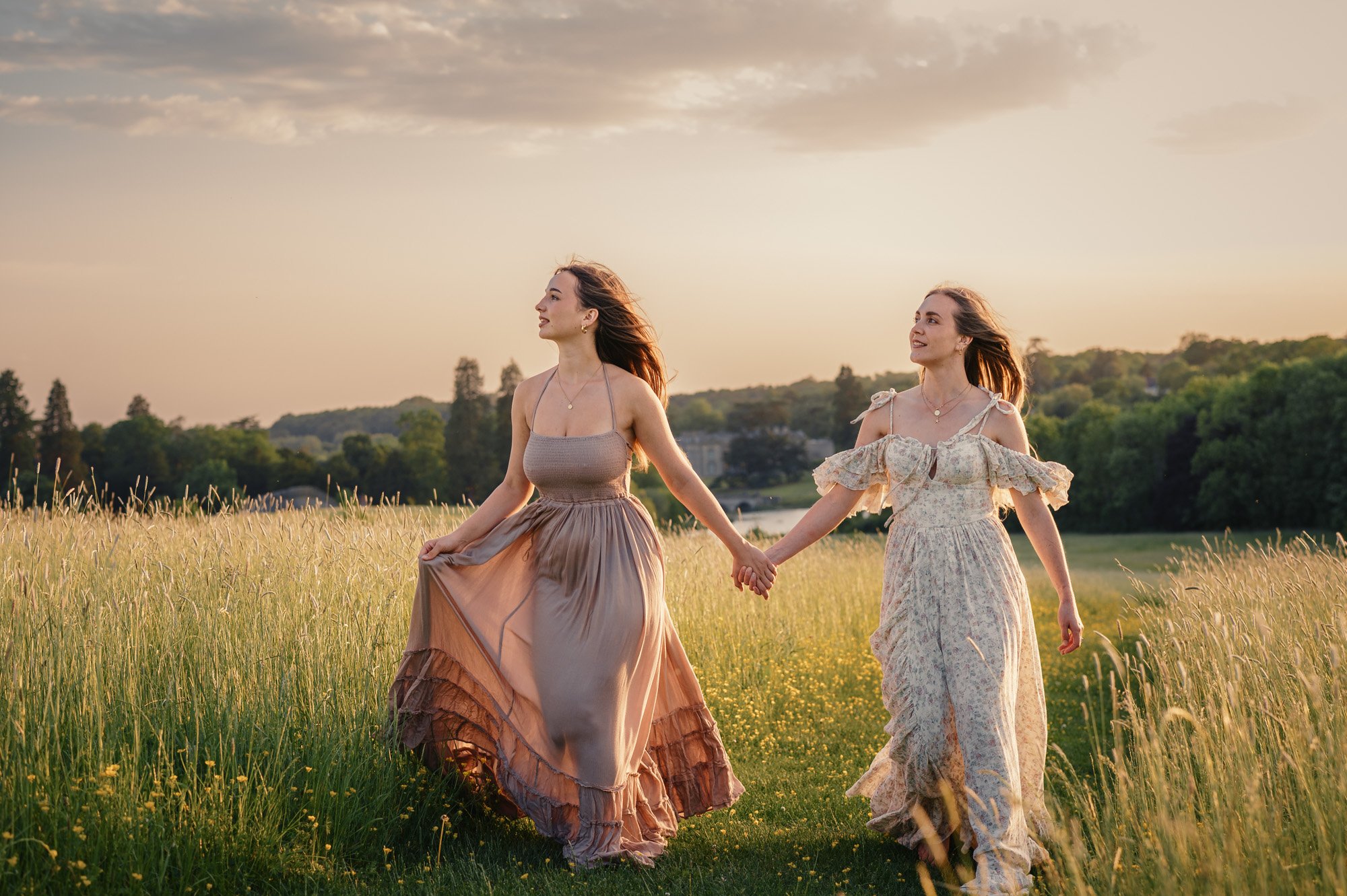 Two women holding hands and walking through a grassy field during sunset, both wearing flowy dresses and smiling, with a backdrop of trees and a cloudy sky.