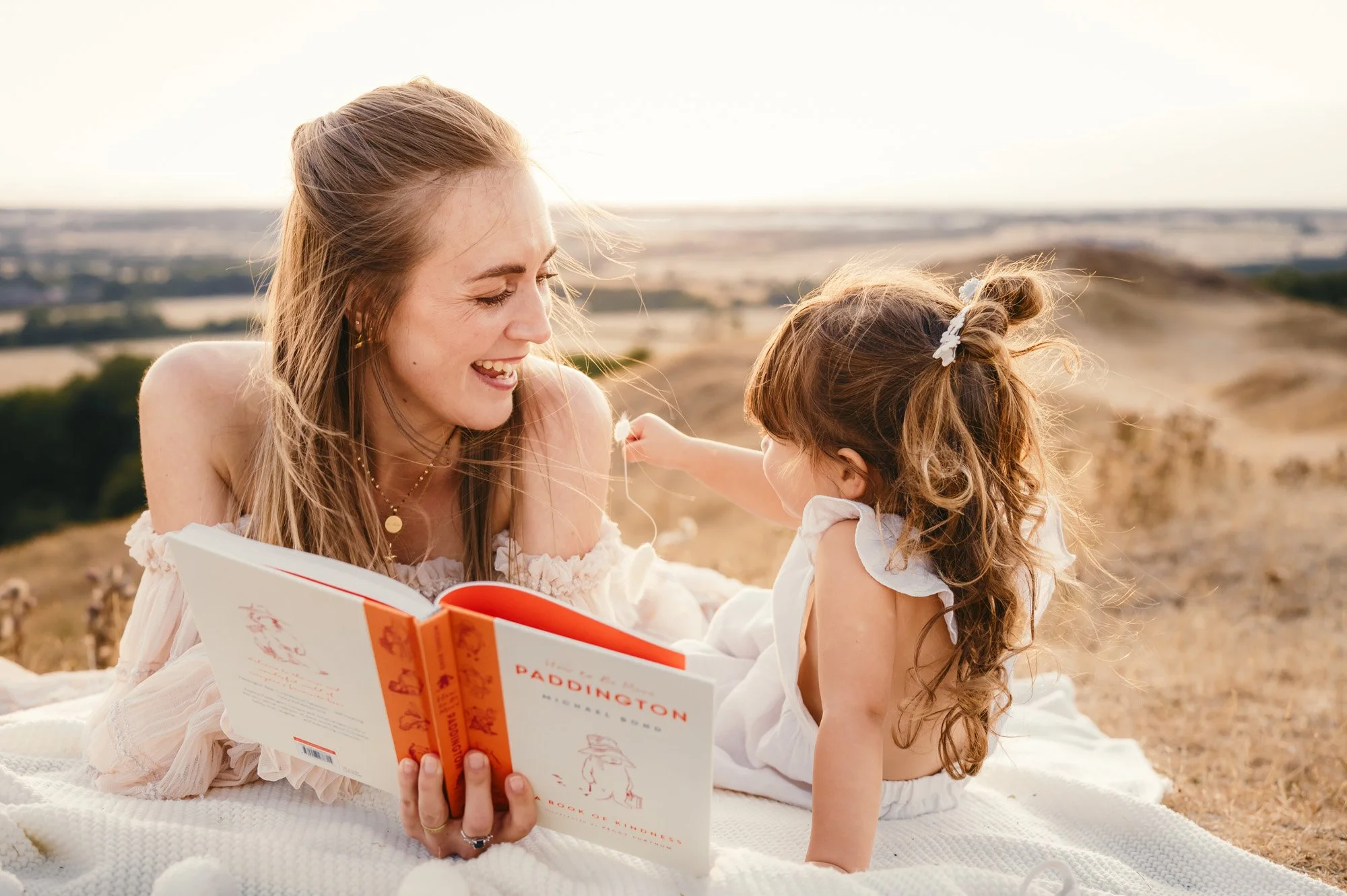 A woman and young girl sitting outdoors on a blanket, reading a book, with a scenic landscape background, both smiling