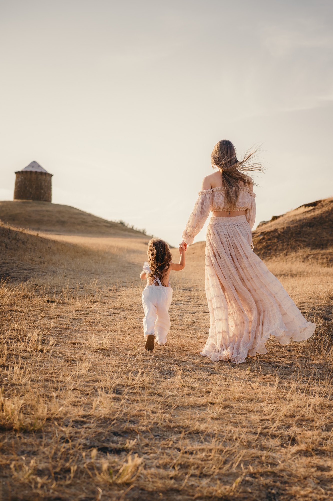 A woman in a flowing, beige, off-the-shoulder dress walking hand-in-hand with a young girl in a white dress in a dry, grassy field at sunset, with a small, round stone building on a hill in the background.