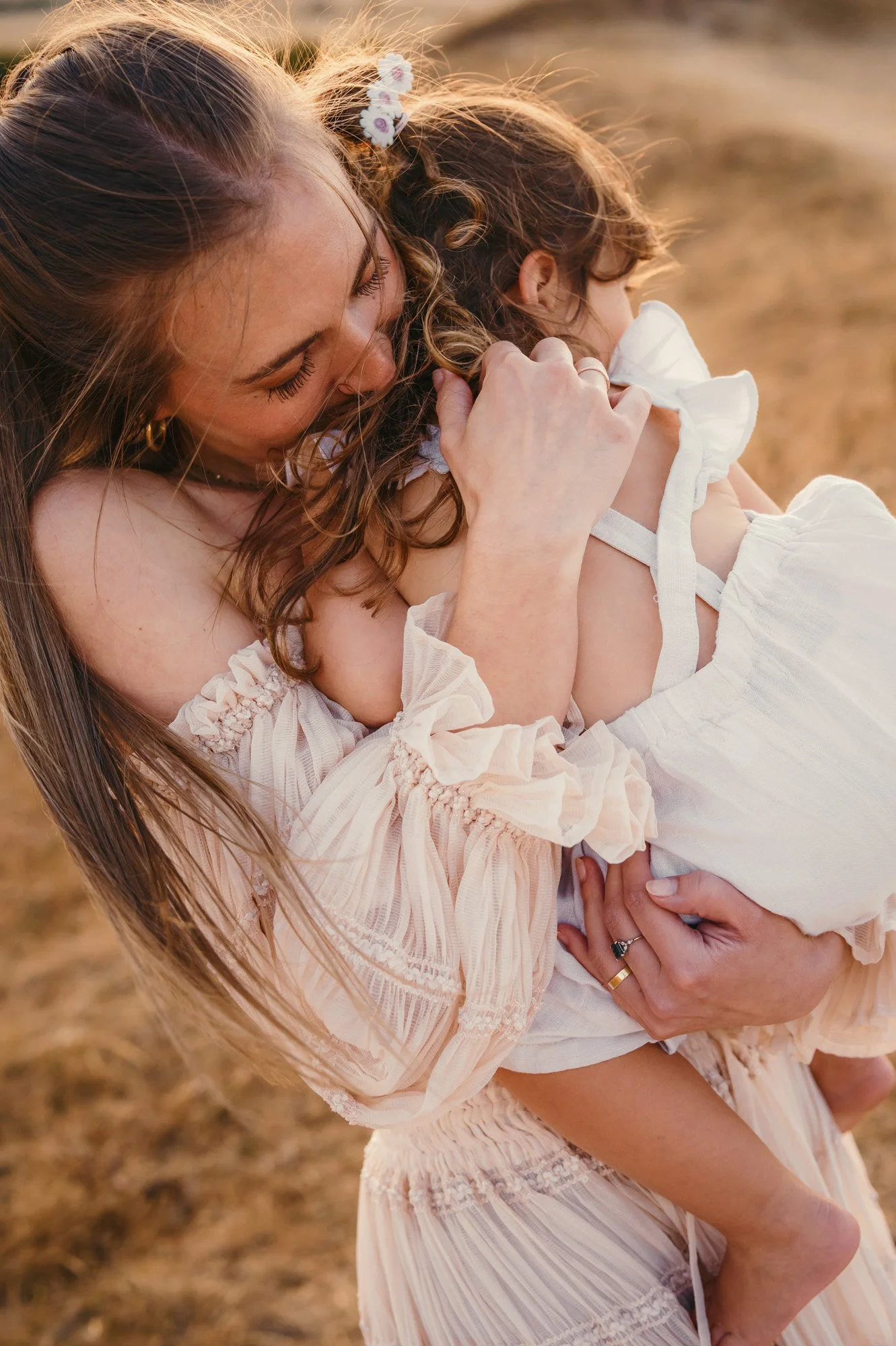 A woman holding a young girl, both with long, curly hair, outdoors during sunset, wearing light-colored, ruffled dresses.