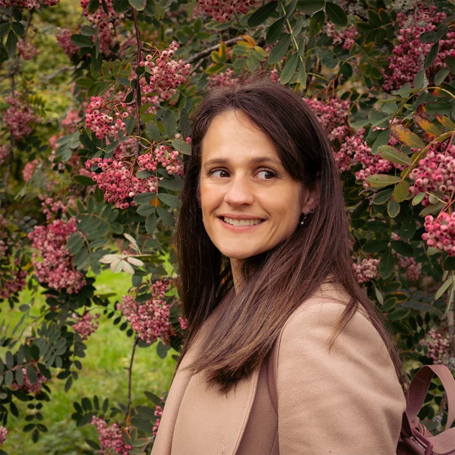 A woman with long brown hair smiling outdoors, standing near a flowering shrub with pink blossoms and green leaves.