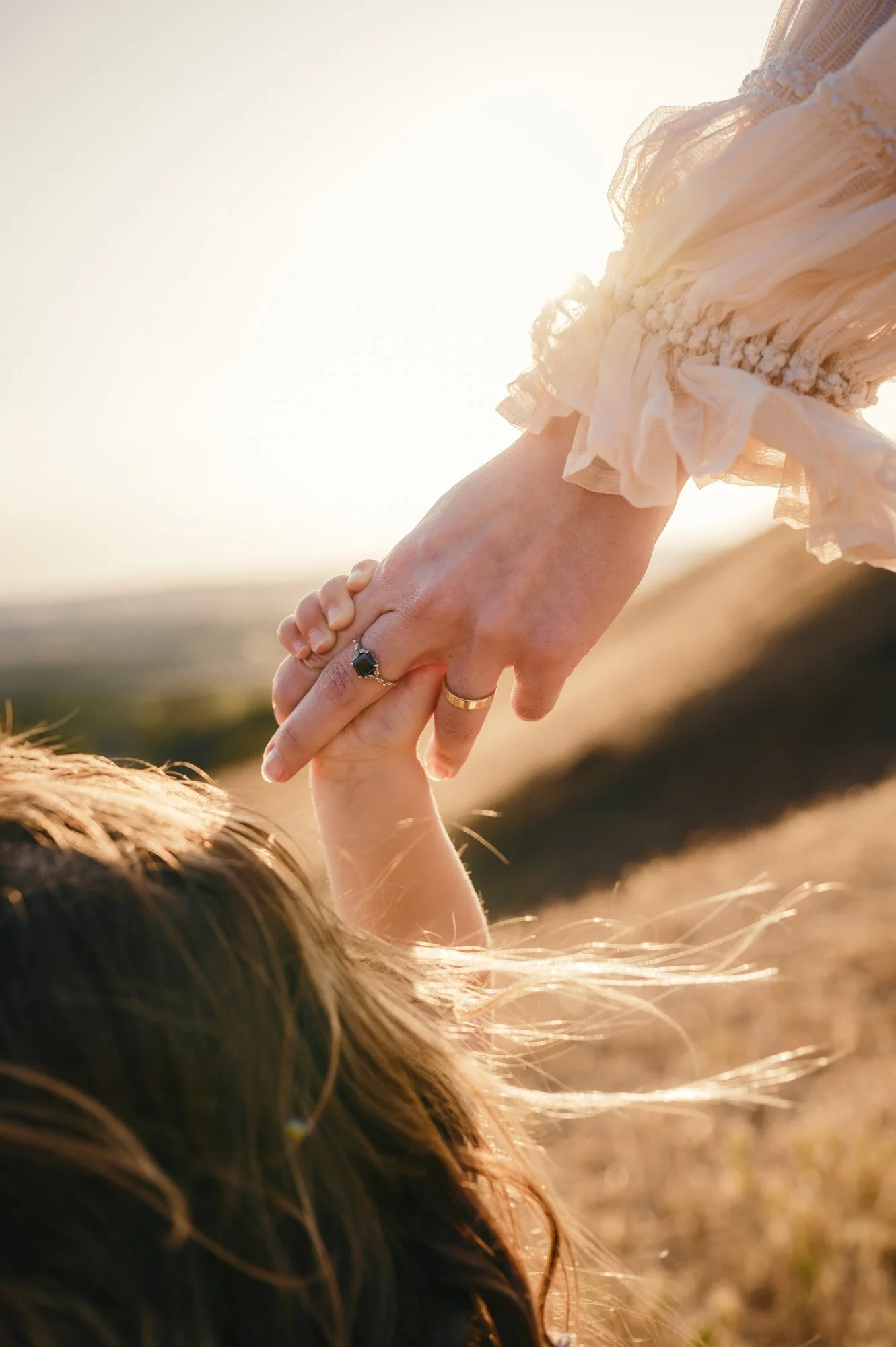 A person holding a child's hand during sunset in a natural outdoor setting, with the child's hair visible in the foreground.