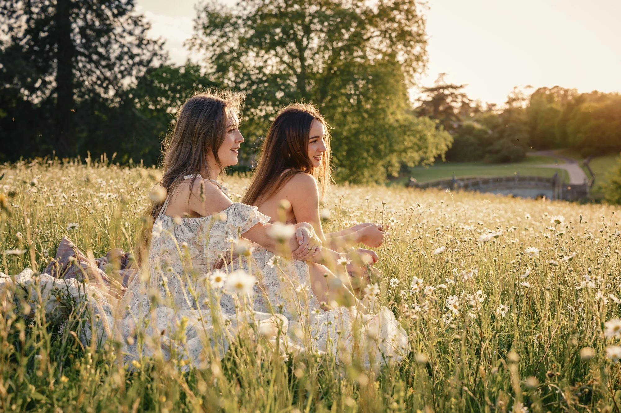 Two young women sitting in a field of daisies during sunset, smiling and enjoying the outdoor scenery with large trees and a bridge in the background.