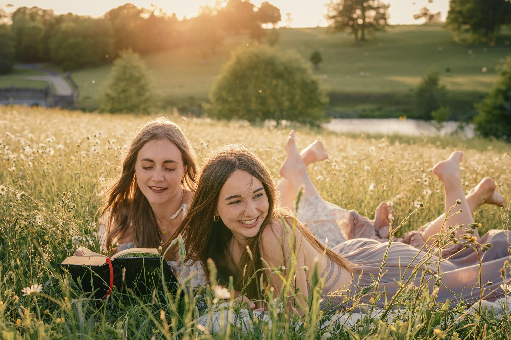 Two young women lying on their stomachs in a field of flowers, reading a book, with trees and a pond in the background during sunset.