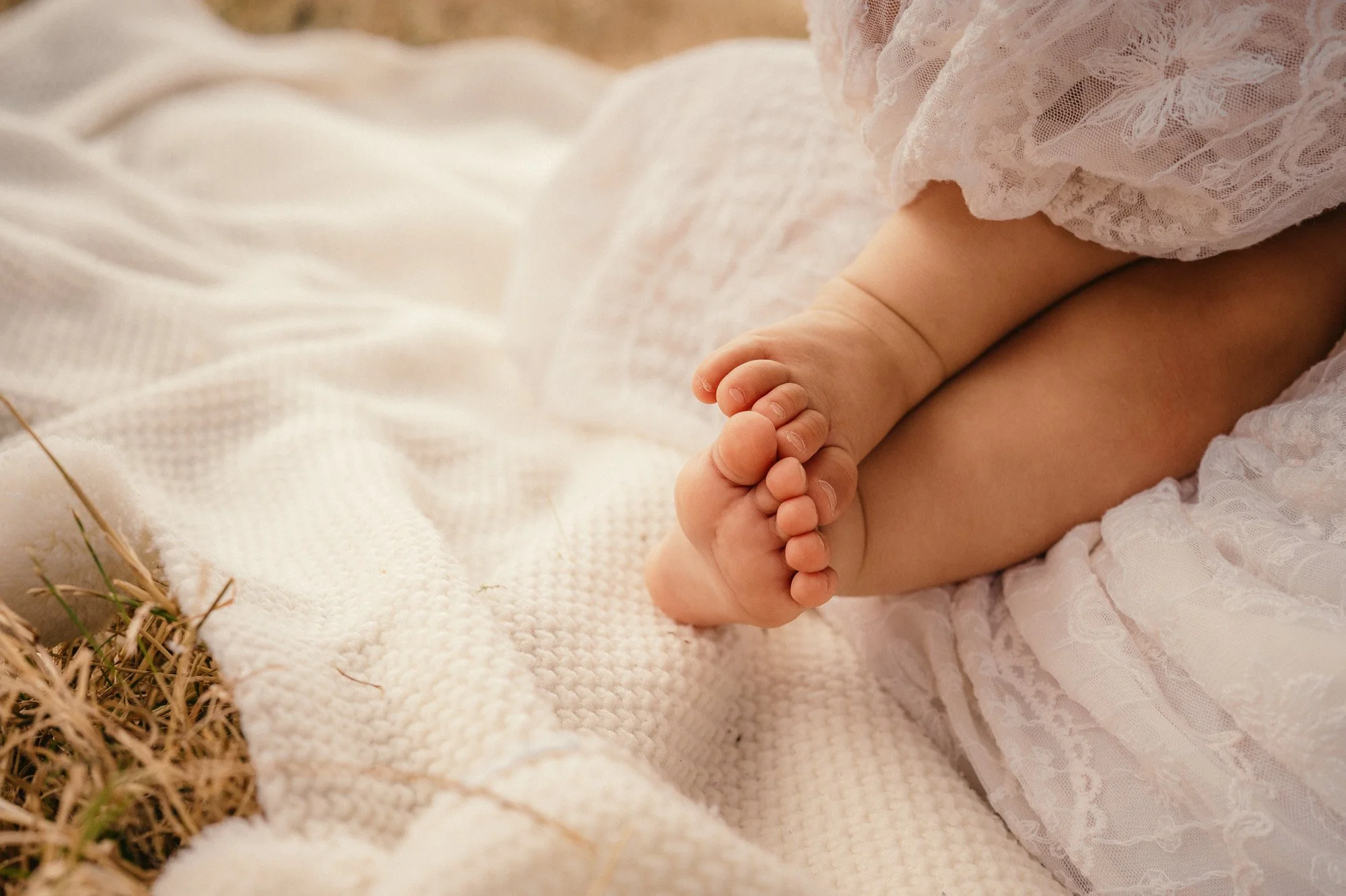Close-up of baby feet and legs wearing lace clothing, lying on a soft white blanket outdoors with some grass in the corner.