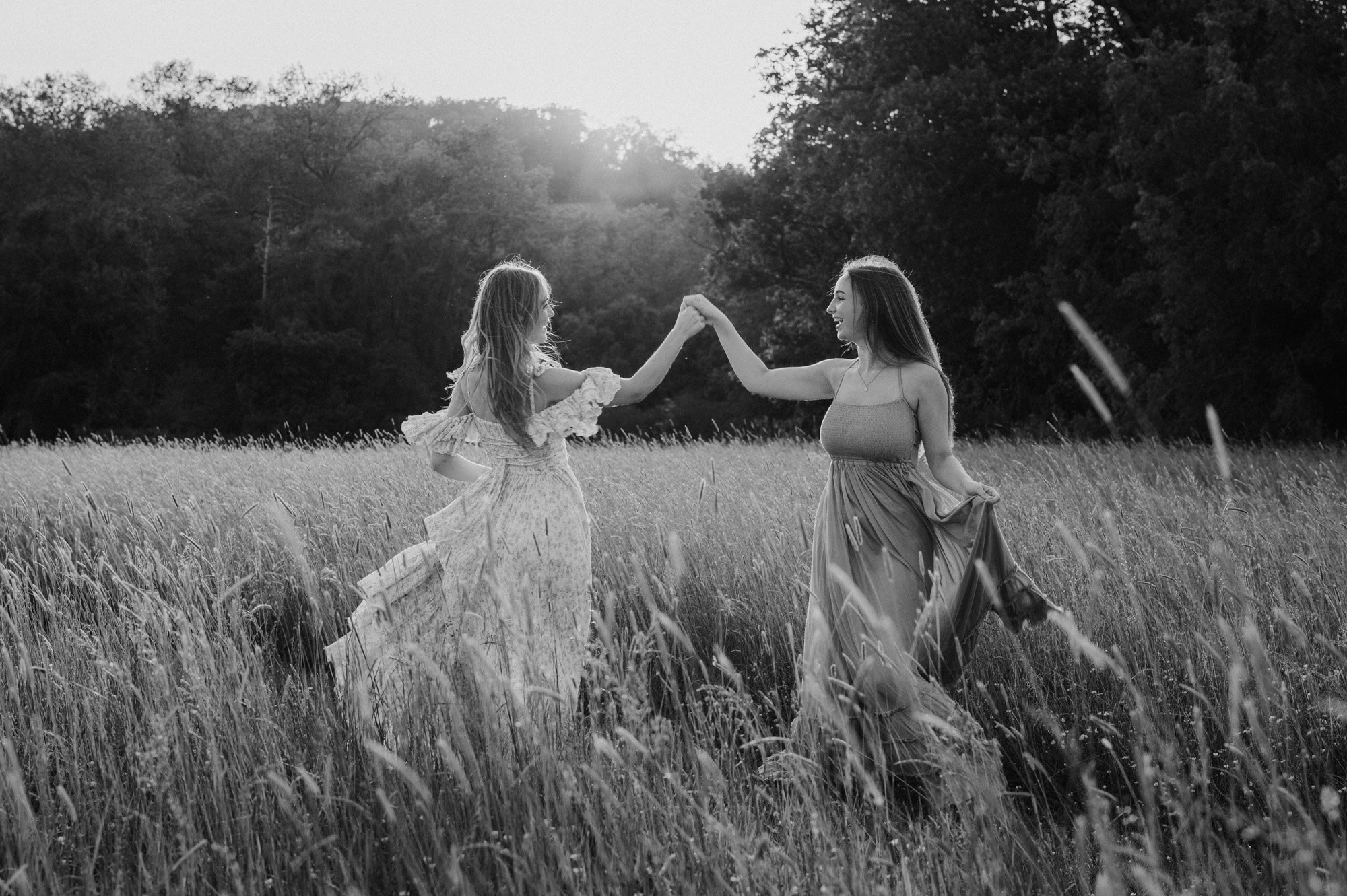 Two women in dresses dancing and smiling in a field of tall grass at sunset, with trees in the background.