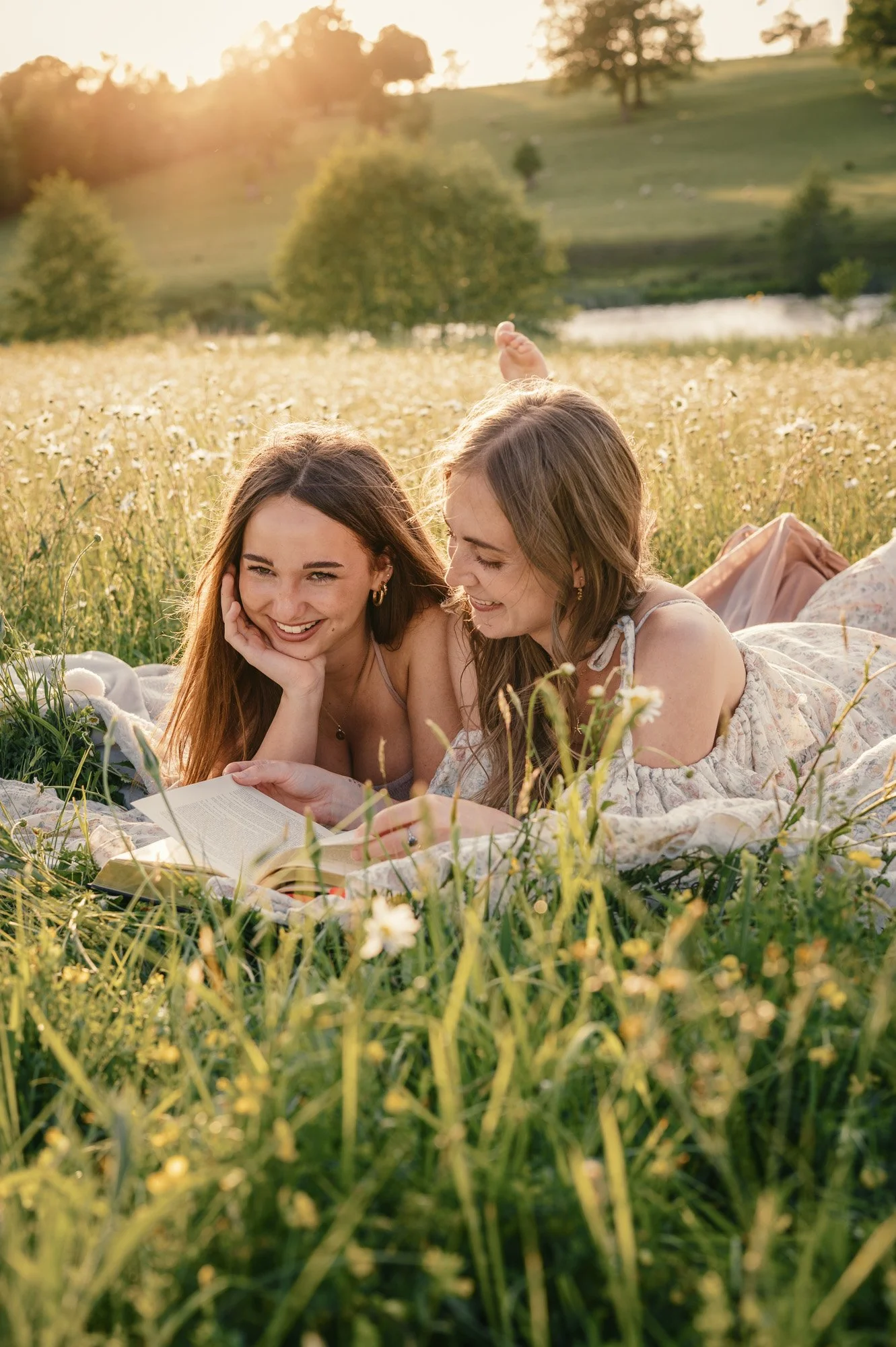 Two young women lying in a grassy field with wildflowers, reading a book and smiling together as the sun sets behind trees in the background.