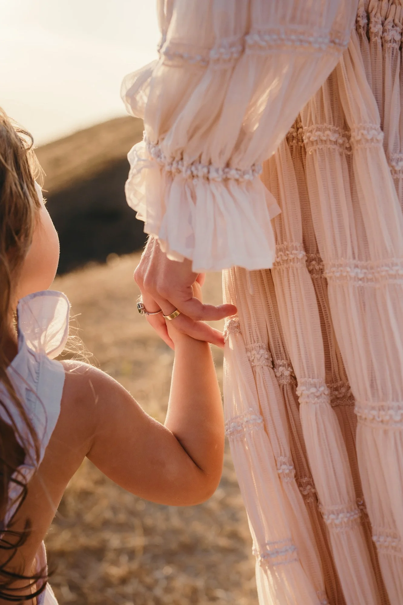 A young girl holding hands with an adult woman, both dressed in light, summery clothing, outdoors during sunset with blurred natural background.