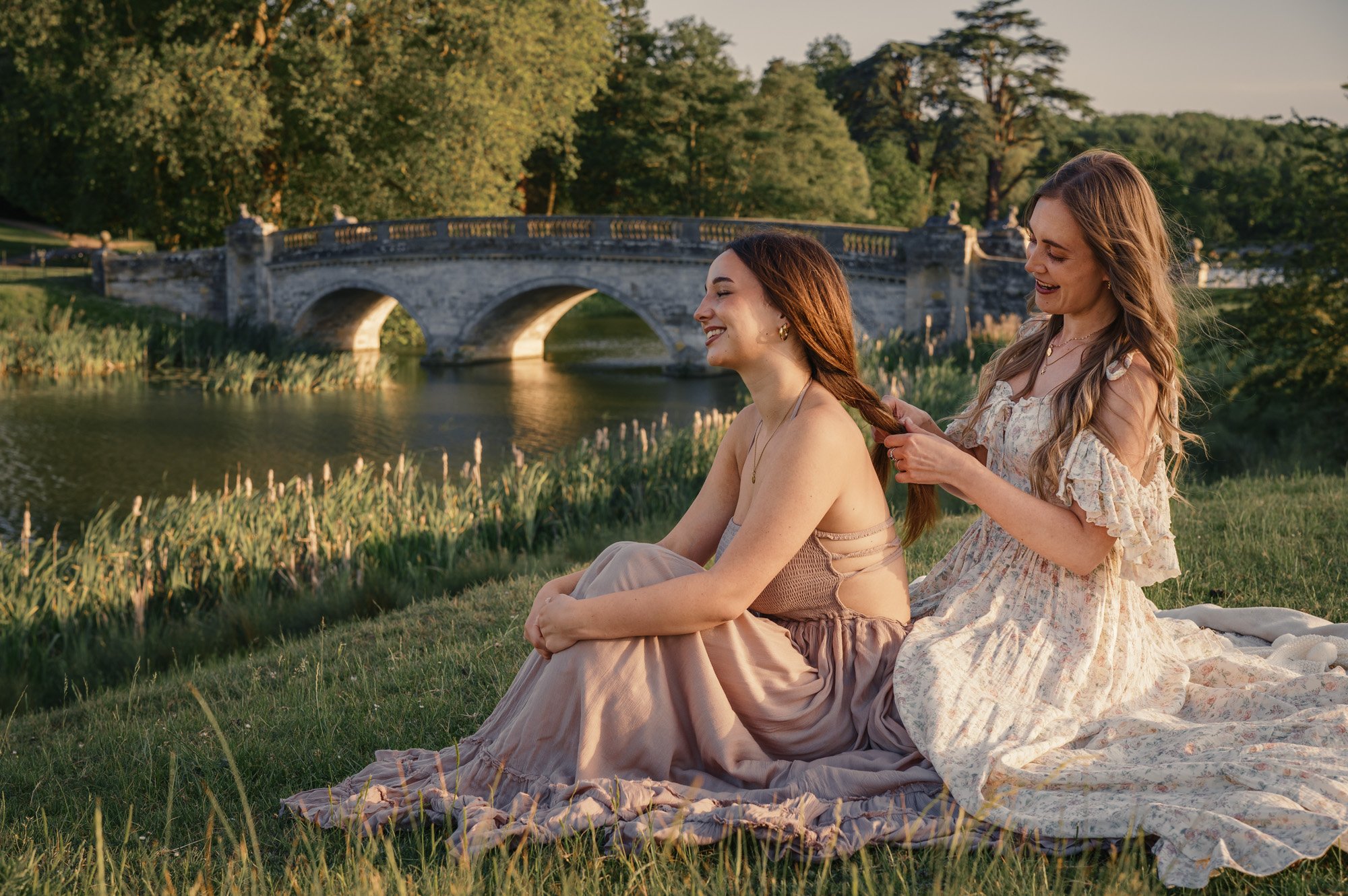 Two women with long hair sitting on a blanket on the grass near a river, with a stone bridge and trees in the background, during sunset.