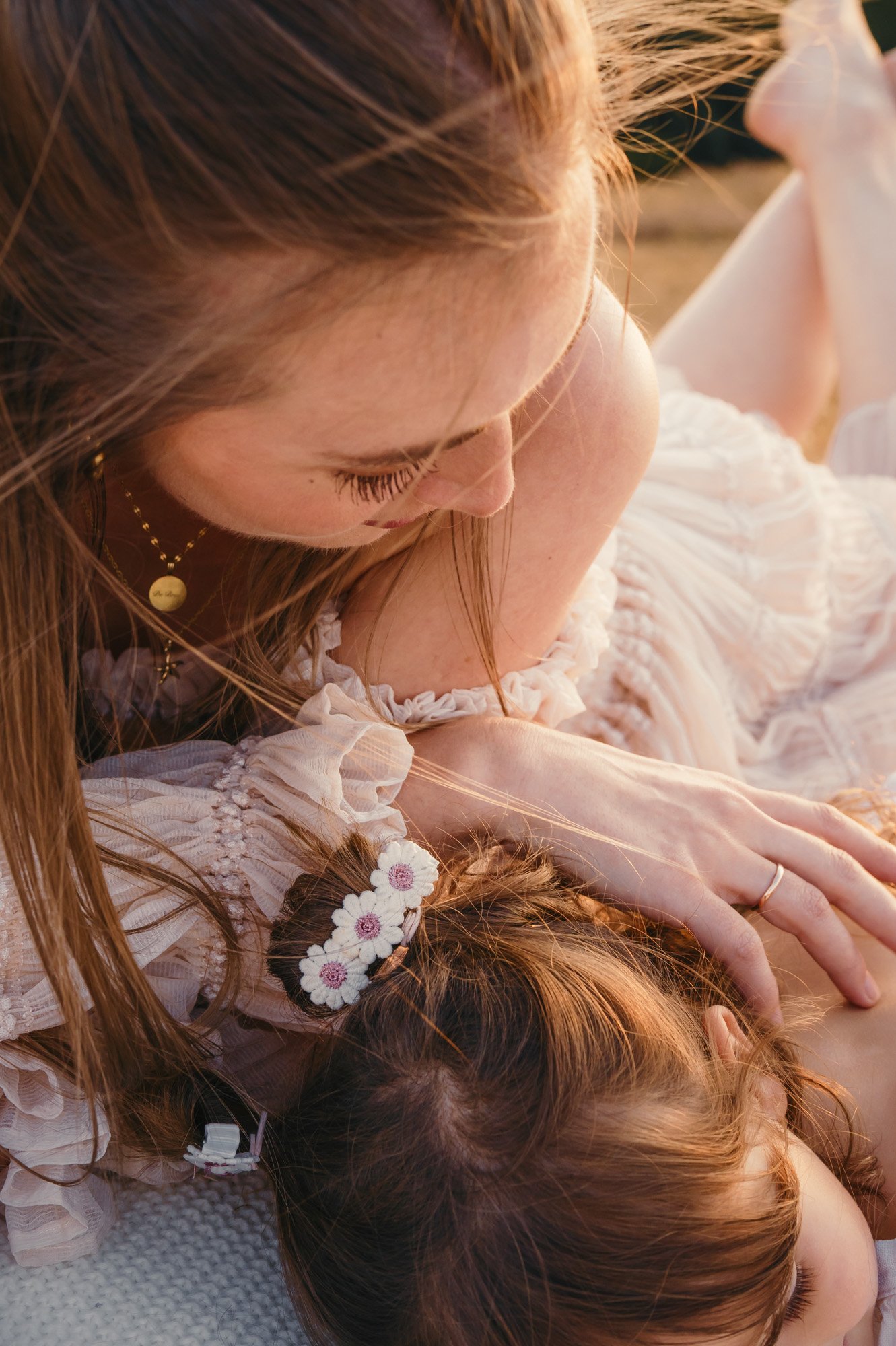 Close-up of a woman with long red hair and a small girl lying on a soft surface. Both have their eyes closed, and the woman appears to be gently resting her hand on the girl's head. The girl has a hairstyle decorated with small white flowers and wear
