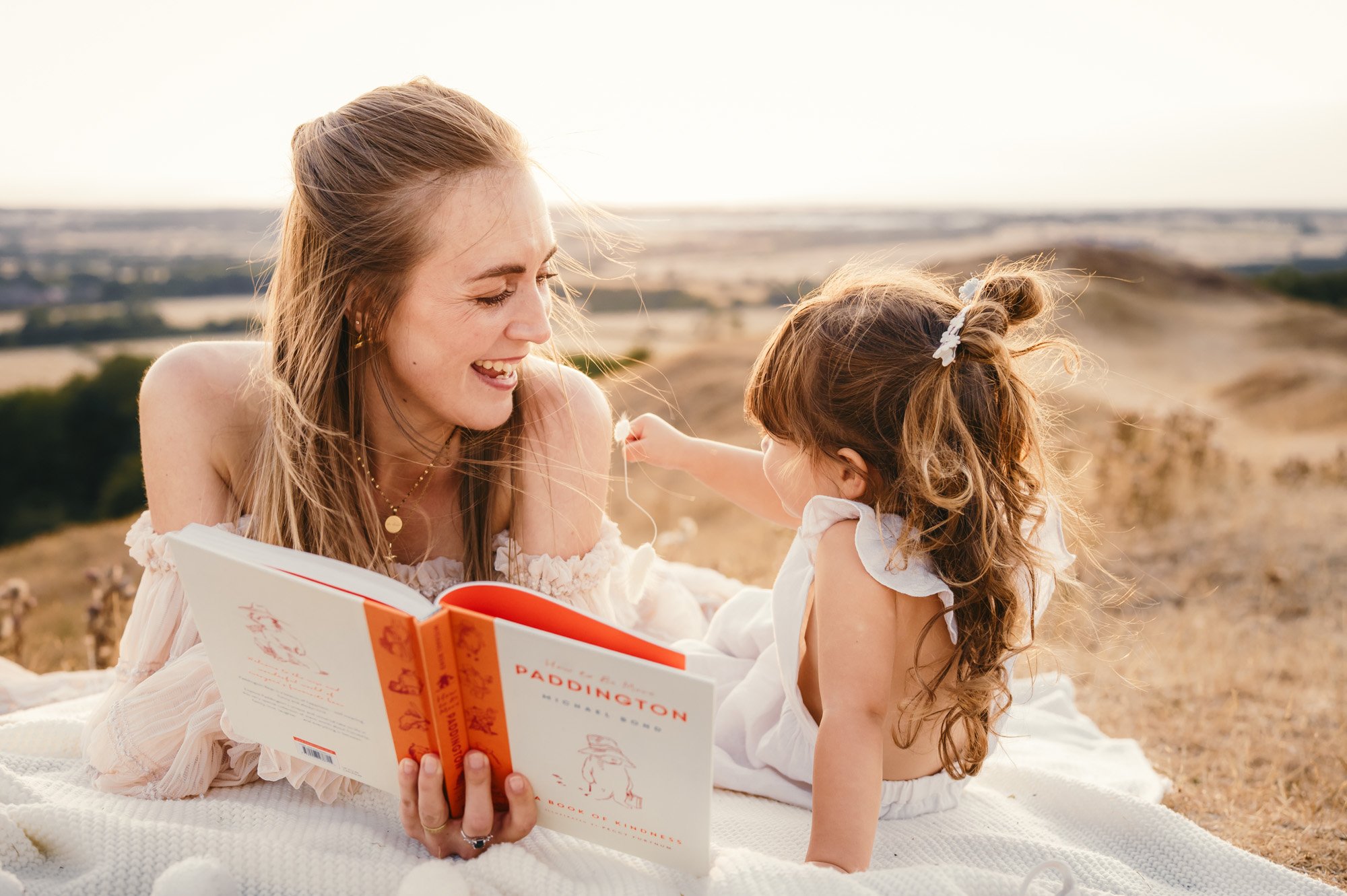 A woman and a young girl sitting outdoors on a blanket, smiling and sharing a moment while the woman reads a book about Paddington Bear. The scene is set on a hill or open field with a blurred landscape in the background, during sunset or late afternoon.