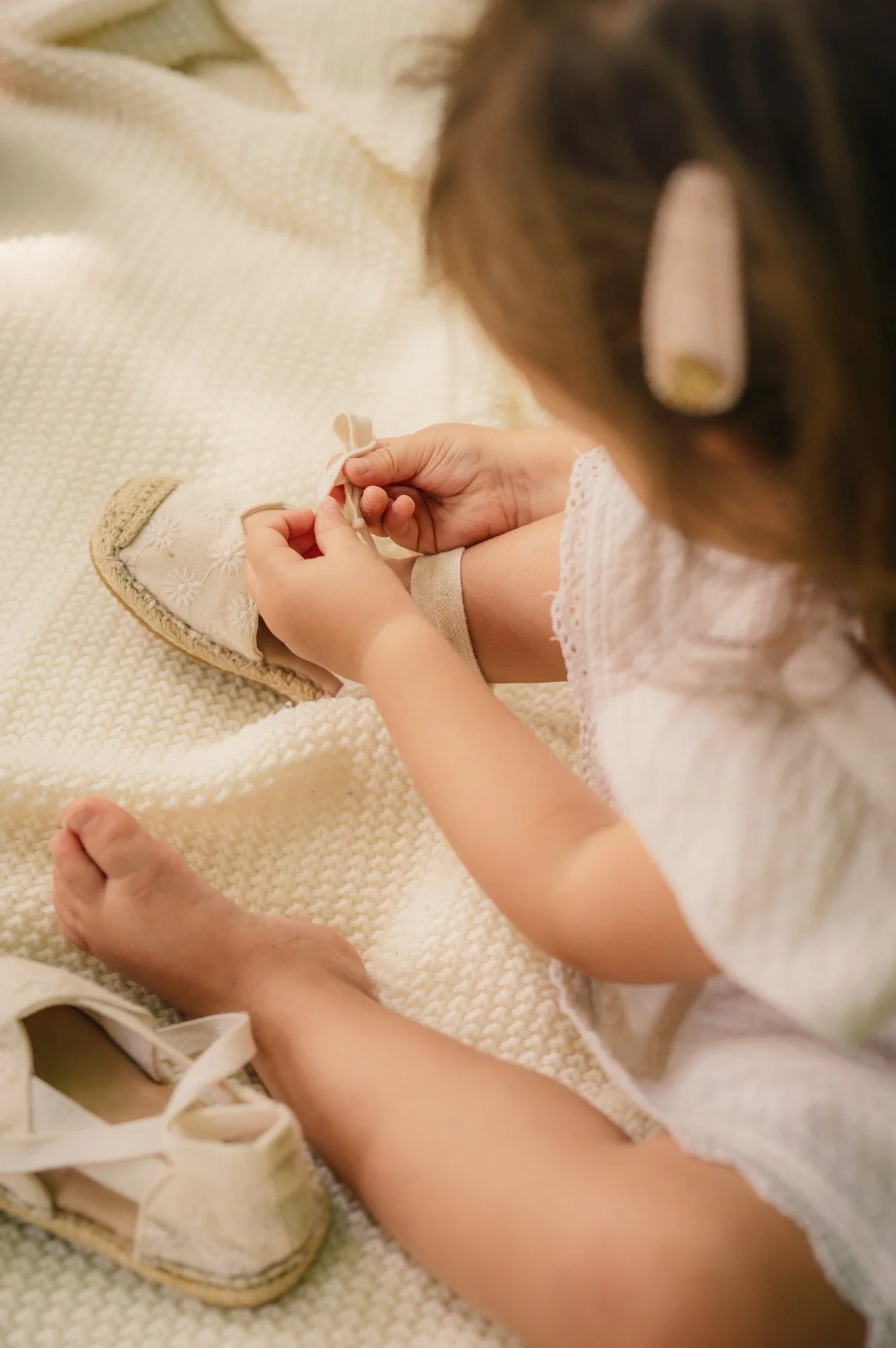 A young girl is sitting on a cream-colored textured blanket, tying the laces of her white embroidered shoe with her hands. Her face is not visible, but her brown hair is partly pinned back with a cloth or hairband. She is wearing a light-colored lace dress.