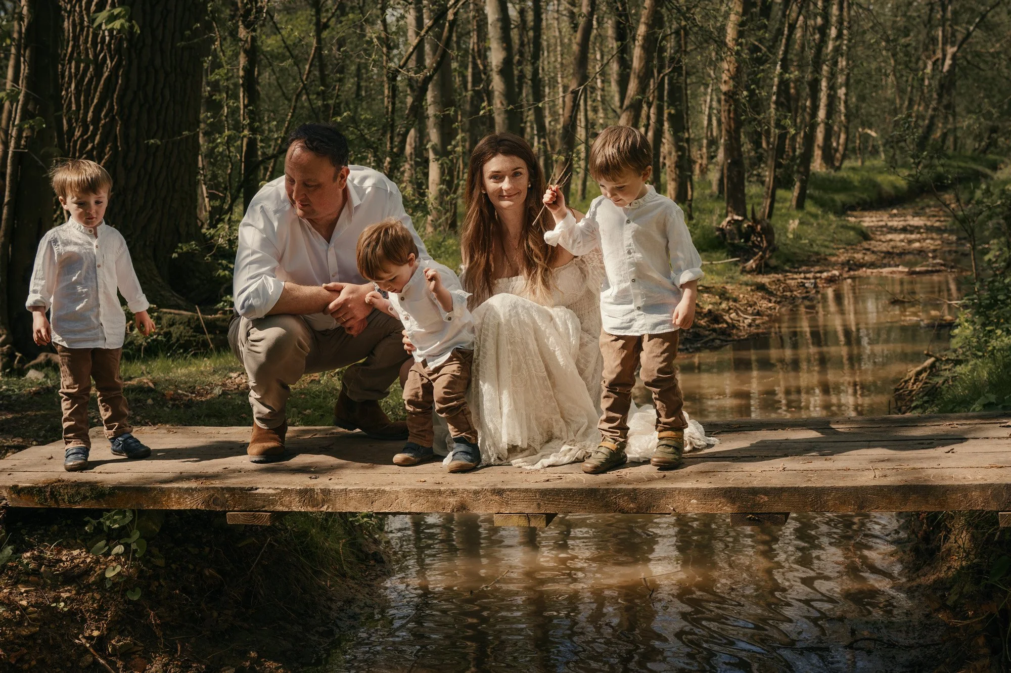 A family of five, including two adults and three young boys, sitting and standing on a wooden dock by a small stream in a forest. The woman wears a long cream dress, and the men and boys wear white shirts and brown pants. They are enjoying a sunny day surrounded by tall trees and green foliage.