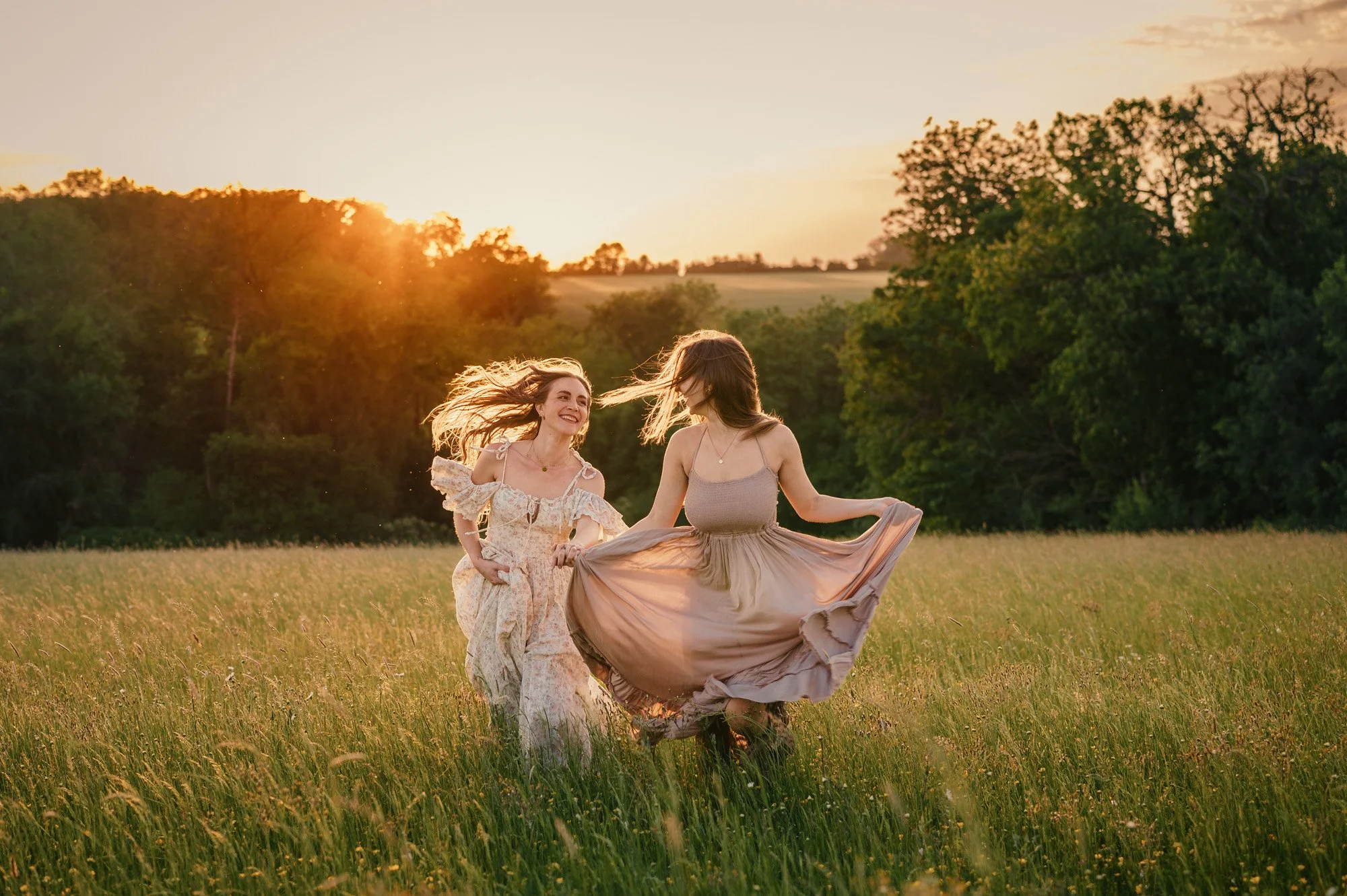 Two women dancing in a grassy field during sunset, wearing flowing dresses and smiling.