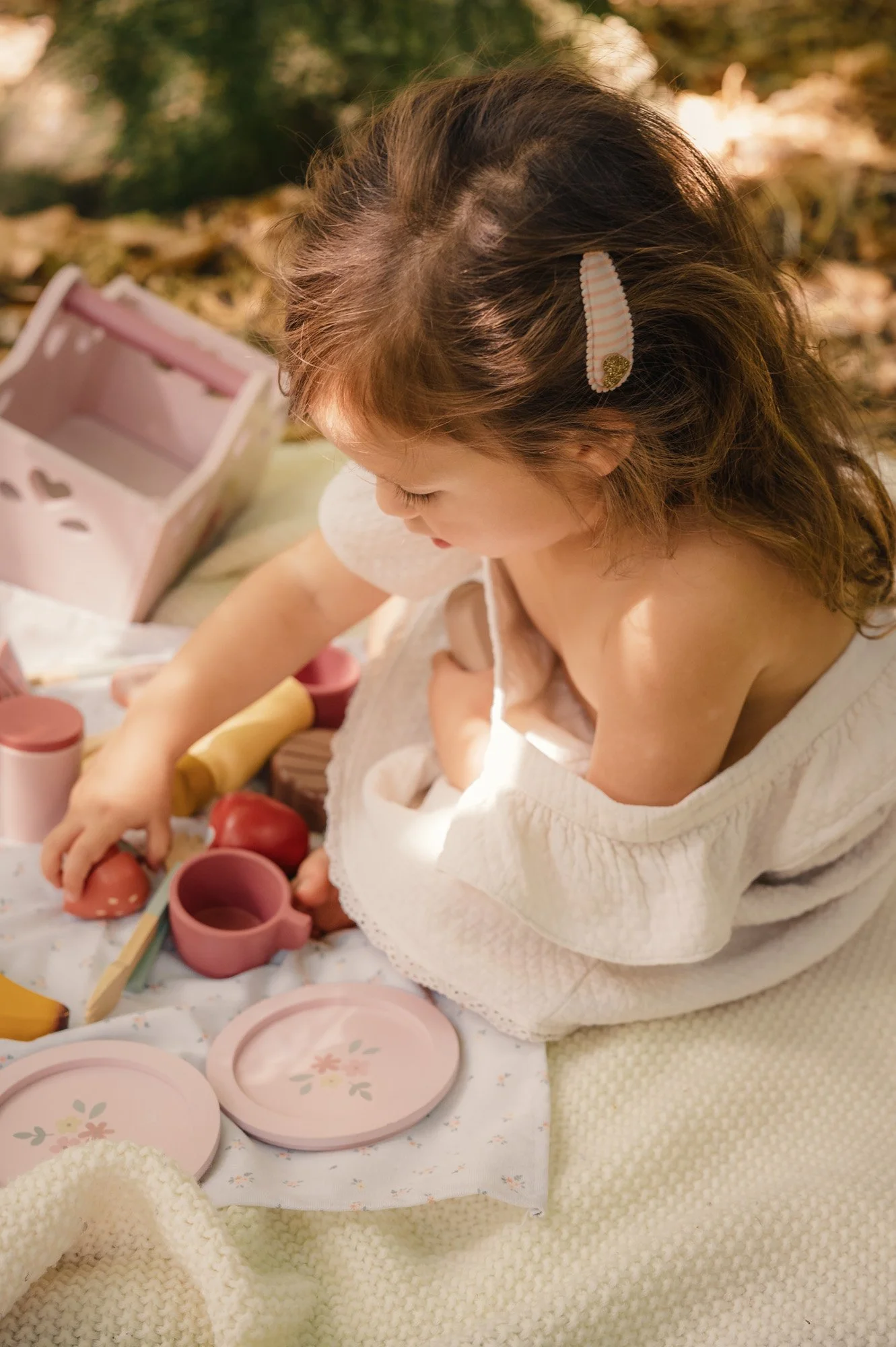 A young girl with brown hair secured with a striped hairclip is sitting on a cozy cream-colored blanket. She is engaged in a pretend tea party with pink and floral patterned dishes, cups, and pretend food items such as apples and rice. The scene is o