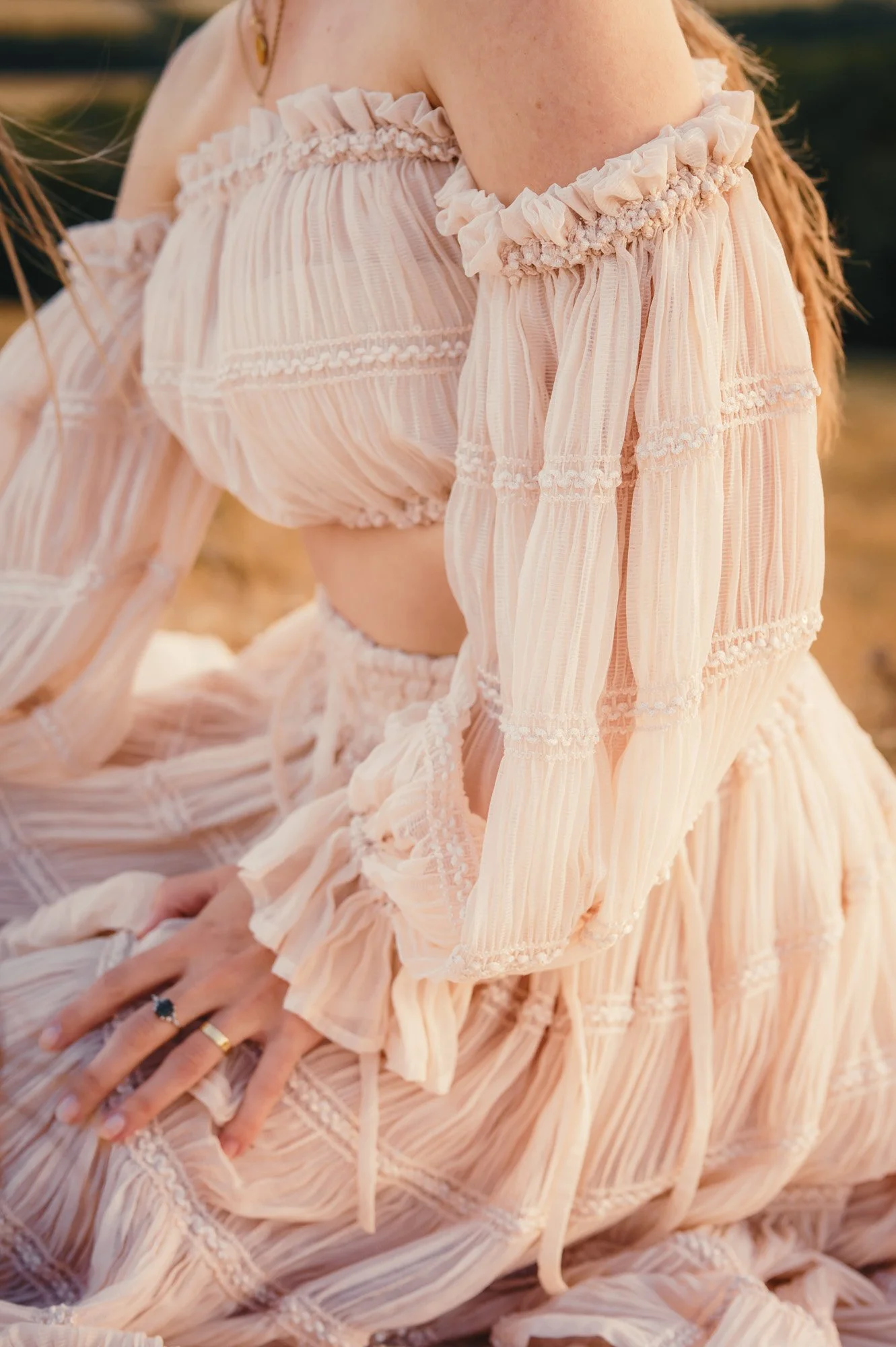 Close-up of a woman wearing a peach-colored, off-the-shoulder dress with ruffles and vertical stripes, sitting outdoors with her hands resting on her lap.