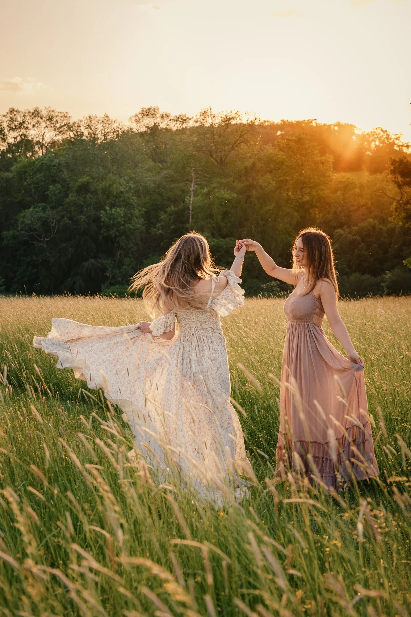 Two women dancing in a sunlit field with tall grass, trees in the background and the sun setting on the horizon.
