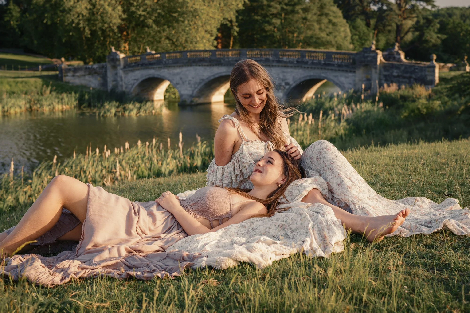 Two young women lying on a blanket in a park with a stone arched bridge and trees in the background, enjoying a sunny day.